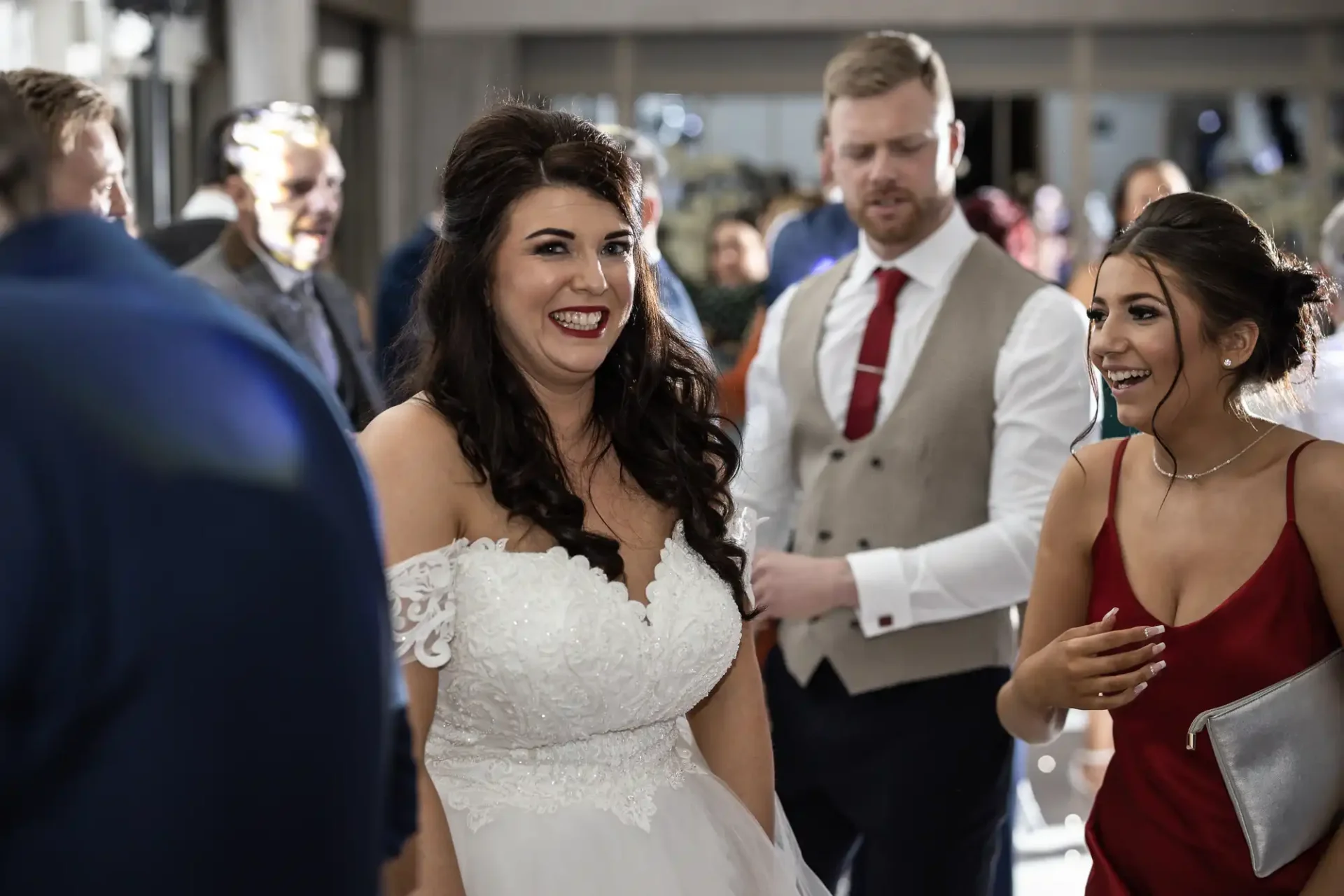 A bride in a white dress laughs with a woman in a green dress at a wedding reception, with a man in a vest and tie walking beside them.
