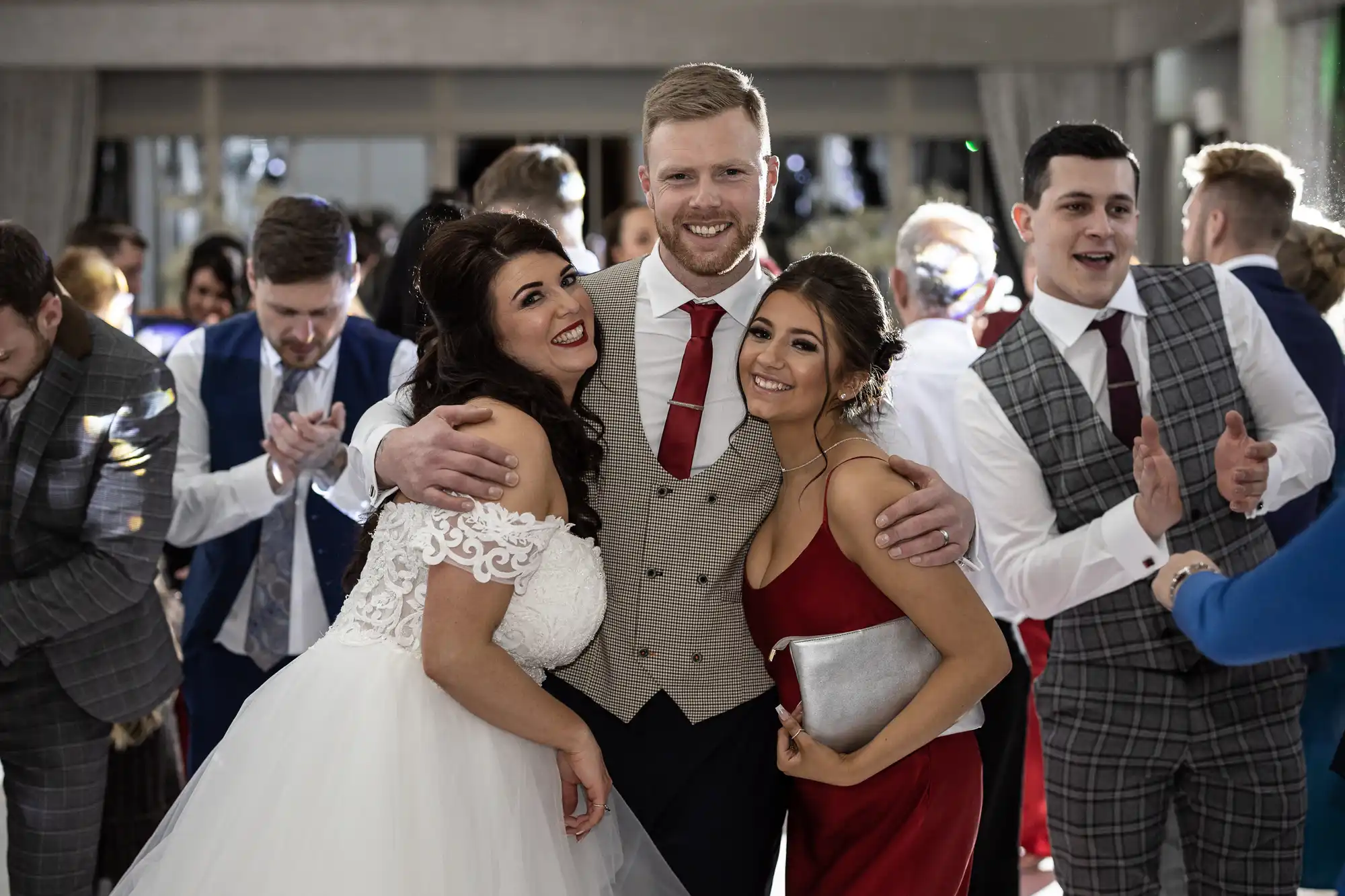 Three people in formal attire, one in a wedding dress, another in a vest with a tie, and the third in a red dress, pose together indoors. Others in the background are clapping and dancing.