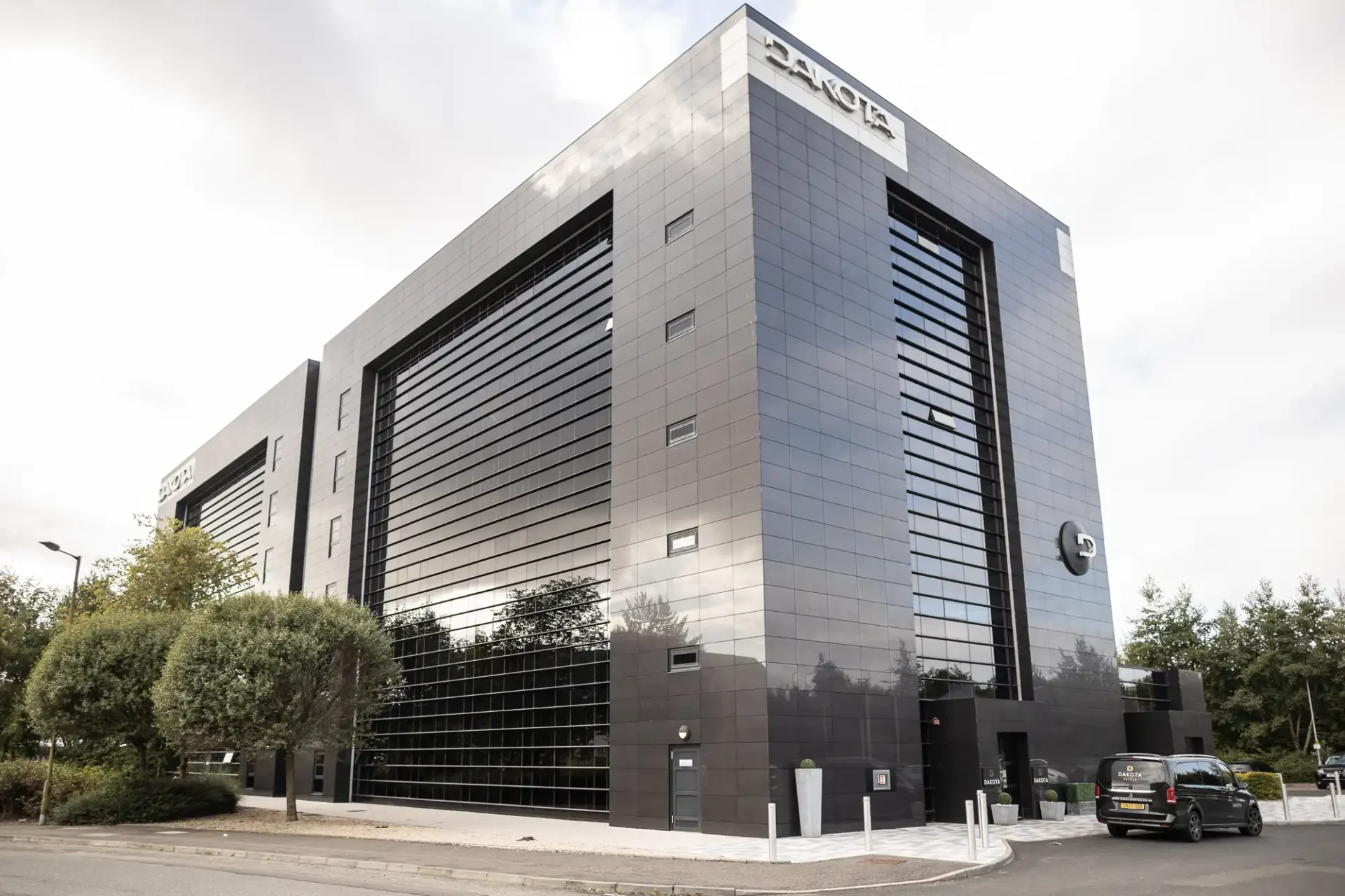 A modern black office building with reflective windows and a logo at the top. A car is parked nearby, and trees are visible in the background.