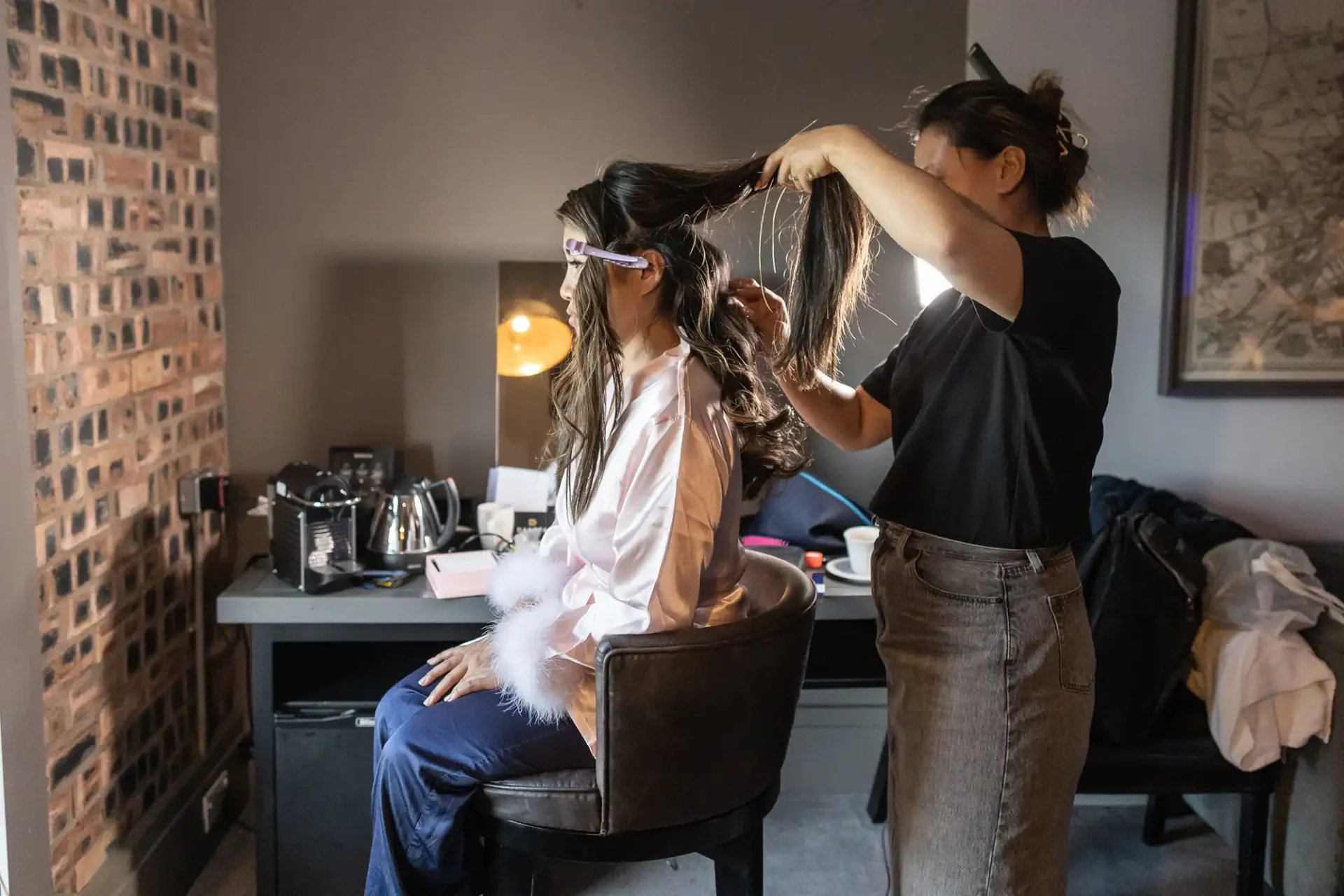 A stylist is curling a seated woman's hair in a room with brick walls, using a hair tool. The woman is wearing a robe and looking at a mirror on the table.