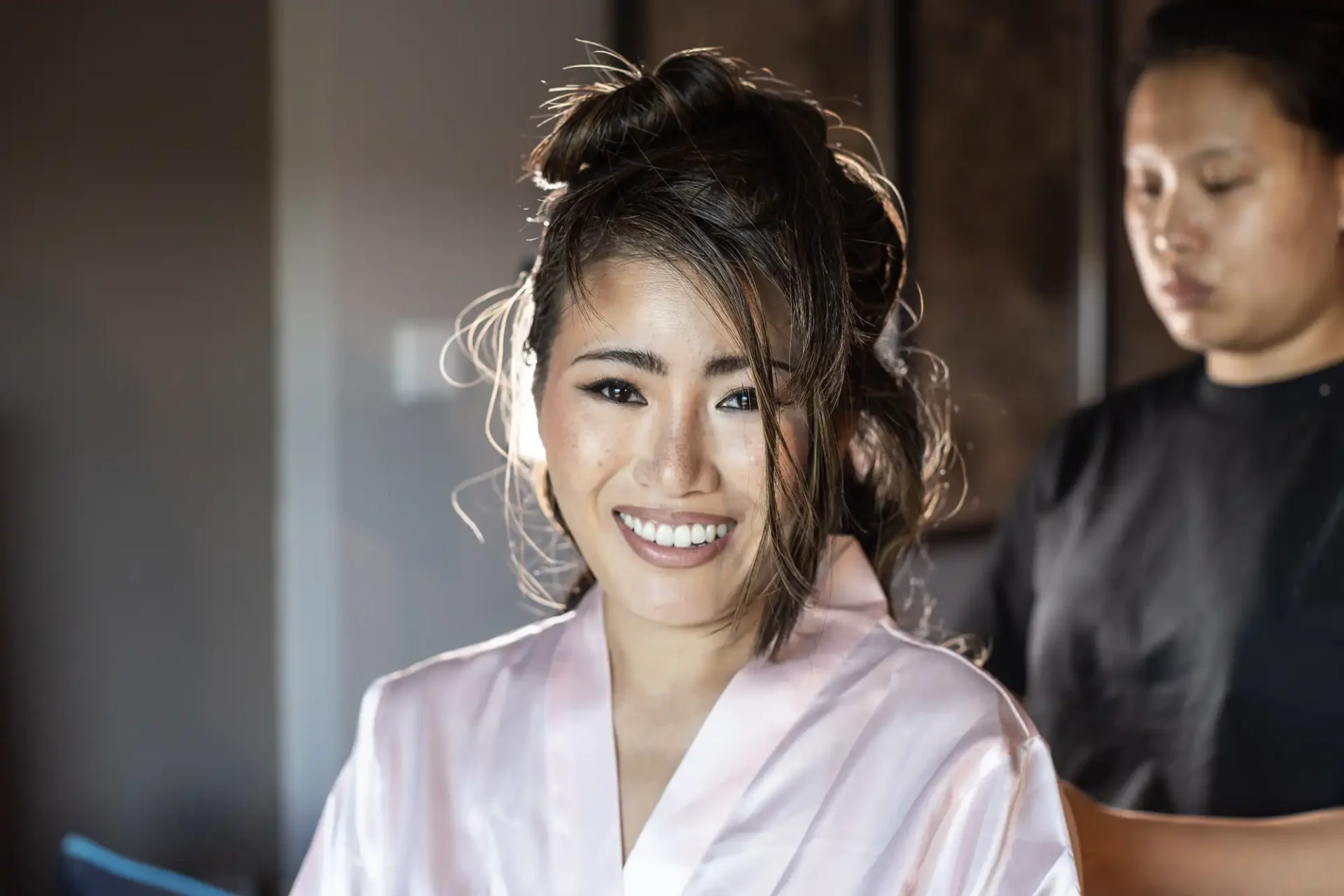 Person in a light robe smiling, with someone styling their hair in the background.