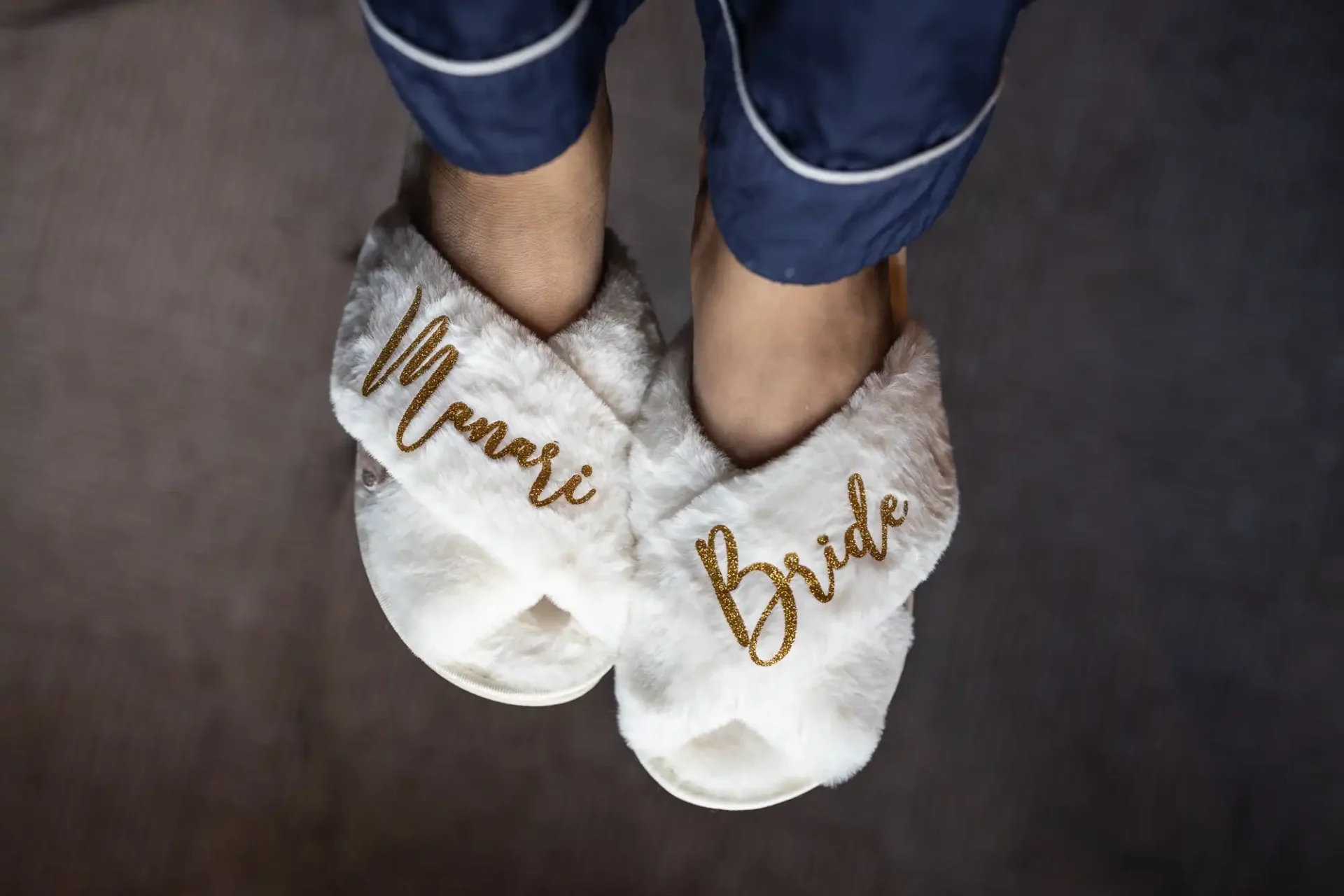 Person wearing fluffy white slippers with gold embroidery, reading "Maman" and "Bride," over a dark floor.