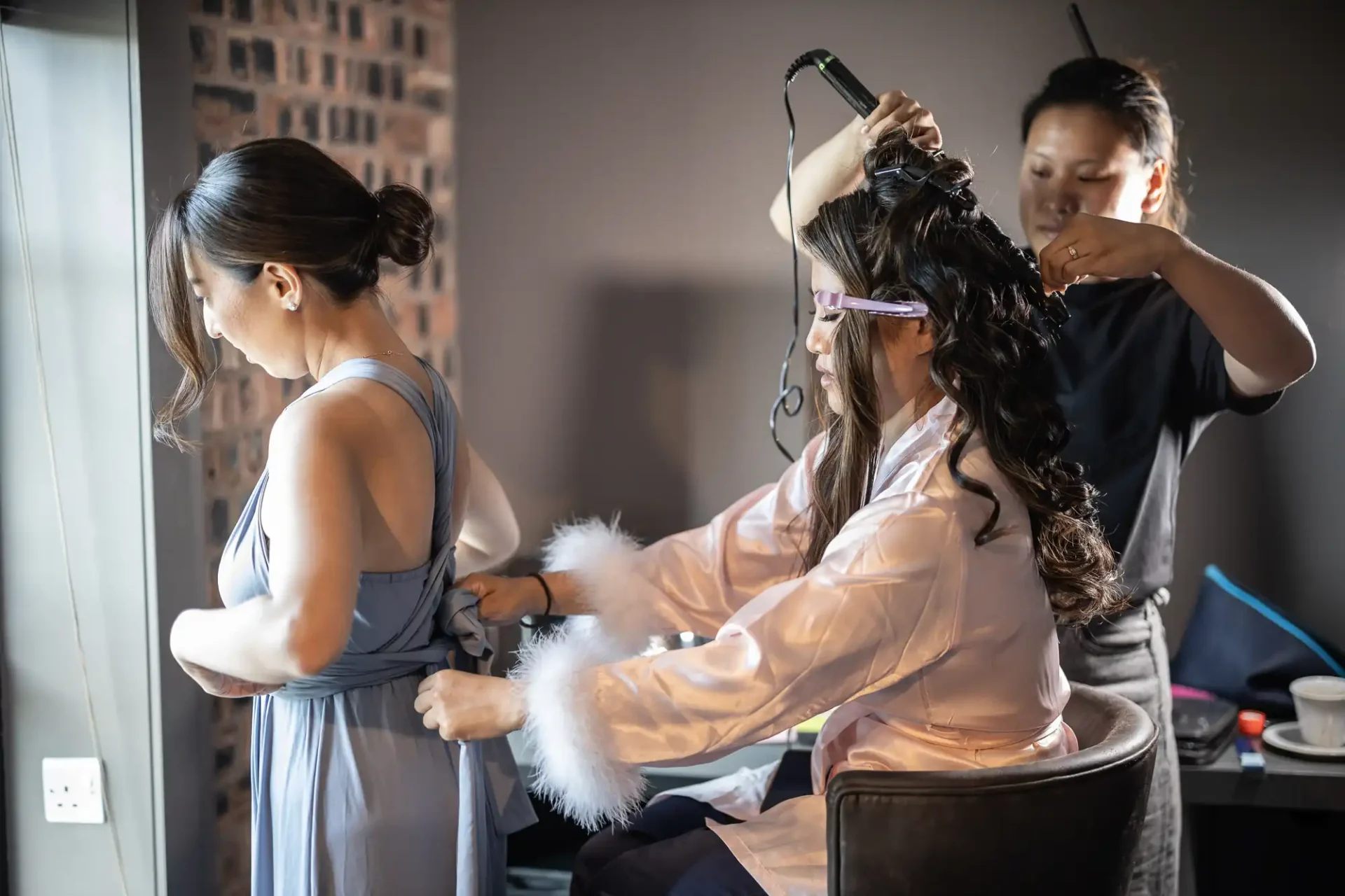 Three women are in a room. One is tying another's dress, who is seated in a robe while the third woman styles her hair with a curling iron.