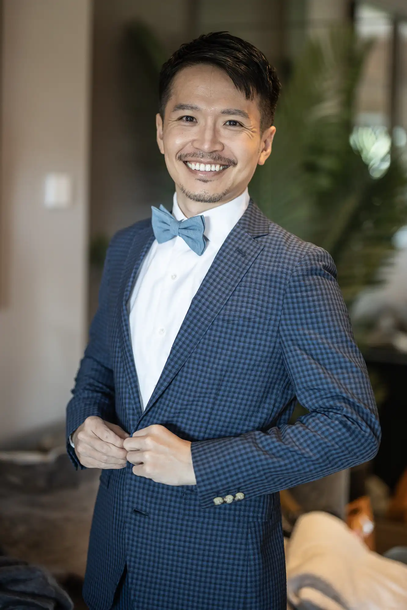 A man in a checkered suit and blue bow tie smiles at the camera, standing in a room with blurred background elements.