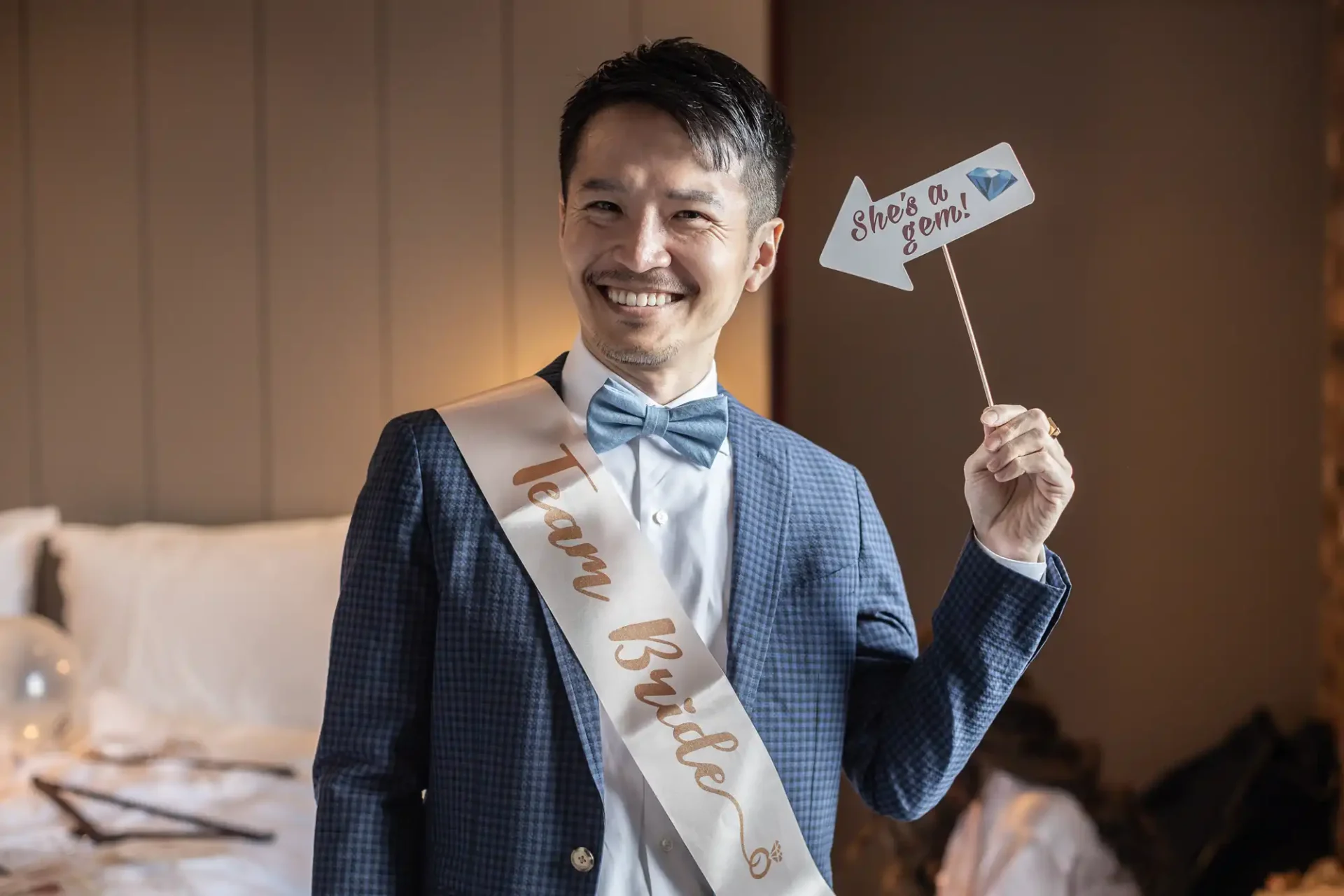 Smiling man wearing a "Team Bride" sash and bow tie, holding a "She's a gem!" sign, in a room with a bed in the background.