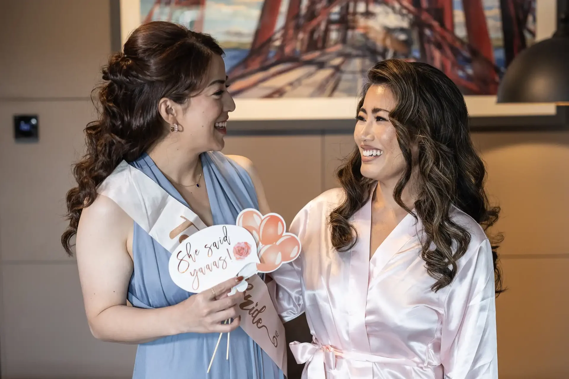 Two women smiling at each other. One in a blue dress holds signs with phrases and balloons, while the other is in a pink robe. A painting of a bridge is in the background.