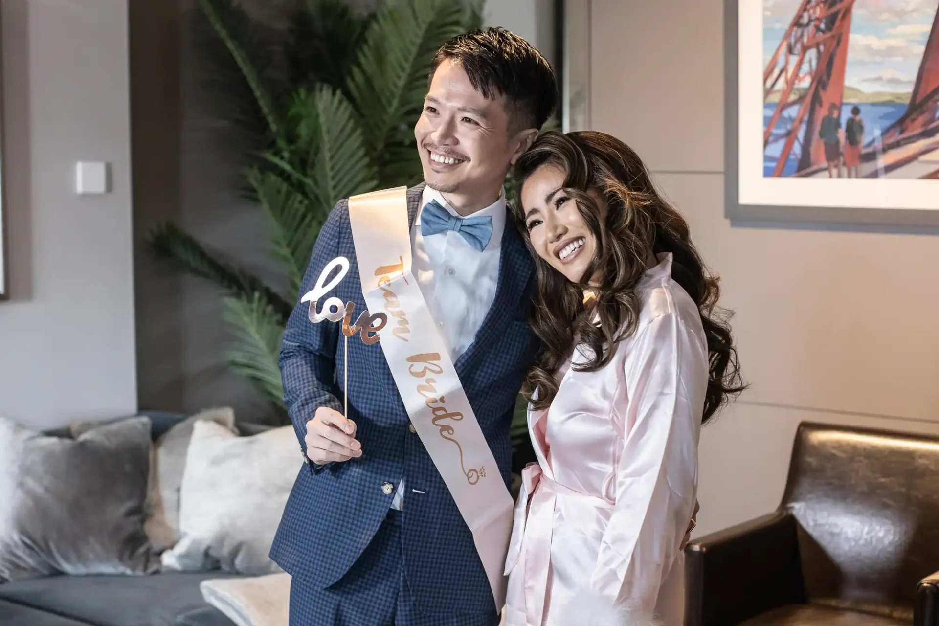 A smiling couple poses indoors. The man wears a "Team Bride" sash and holds a "love" sign. They are dressed in formal attire with a sofa and artwork visible in the background.