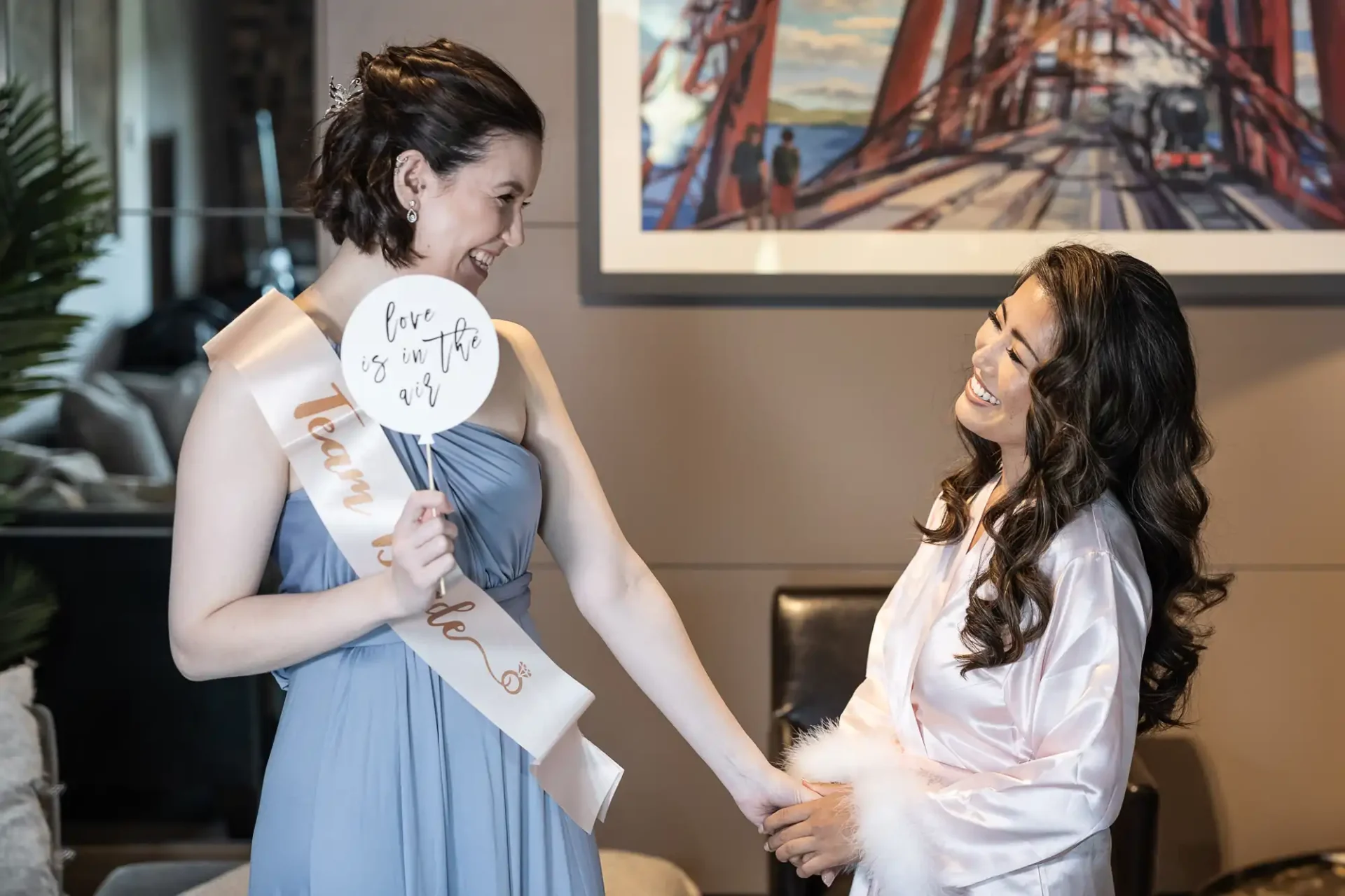 Two women smiling at each other, one wearing a gray dress and holding bride-themed props, the other in a white satin outfit.