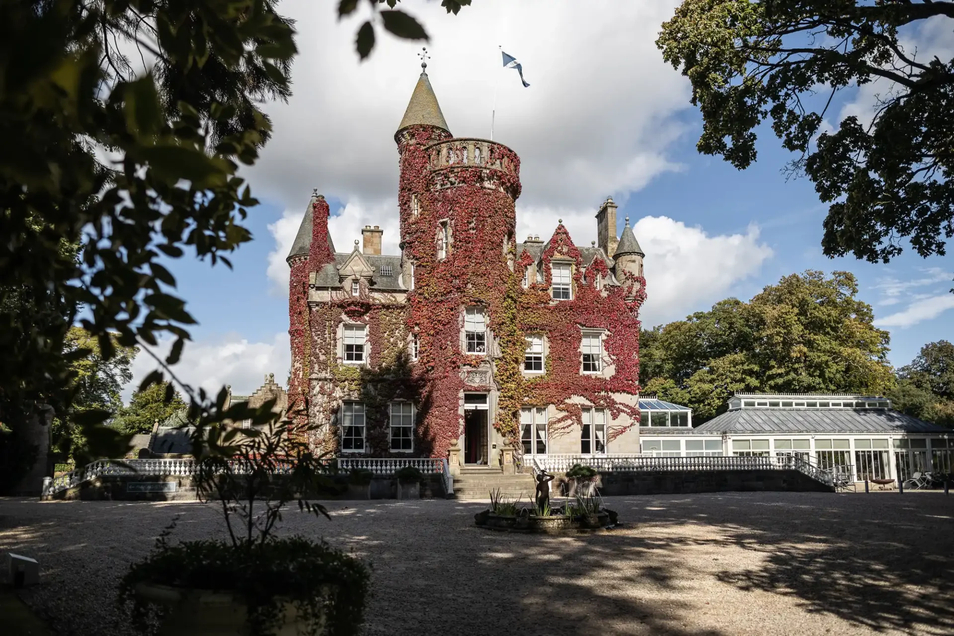 A historic castle covered in red ivy stands against a partly cloudy sky, with a courtyard and greenery around.