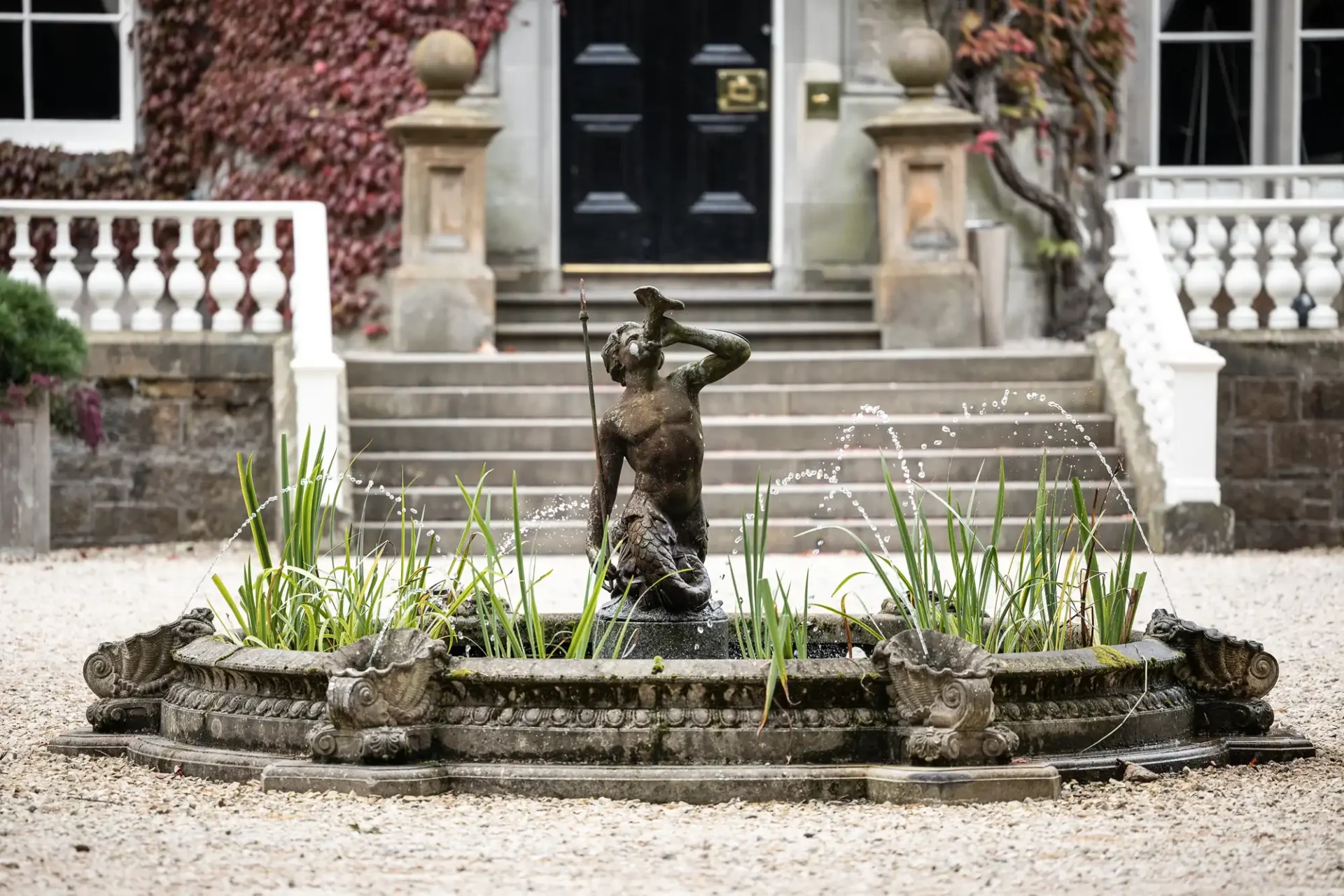 A stone fountain with a statue of a man holding a spear is set in a courtyard with steps and a building in the background.