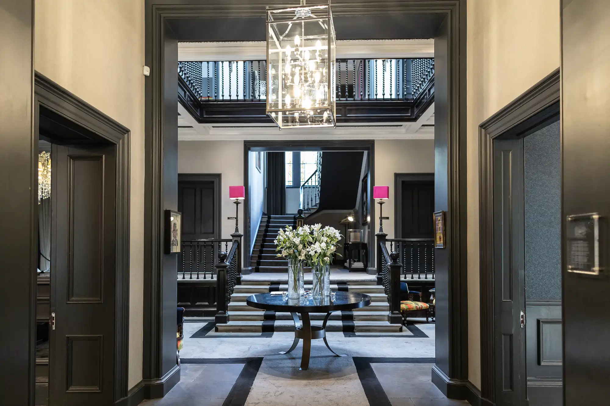Spacious interior with a round table and flowers, chandelier above, staircase in the background, and dark doors on either side.