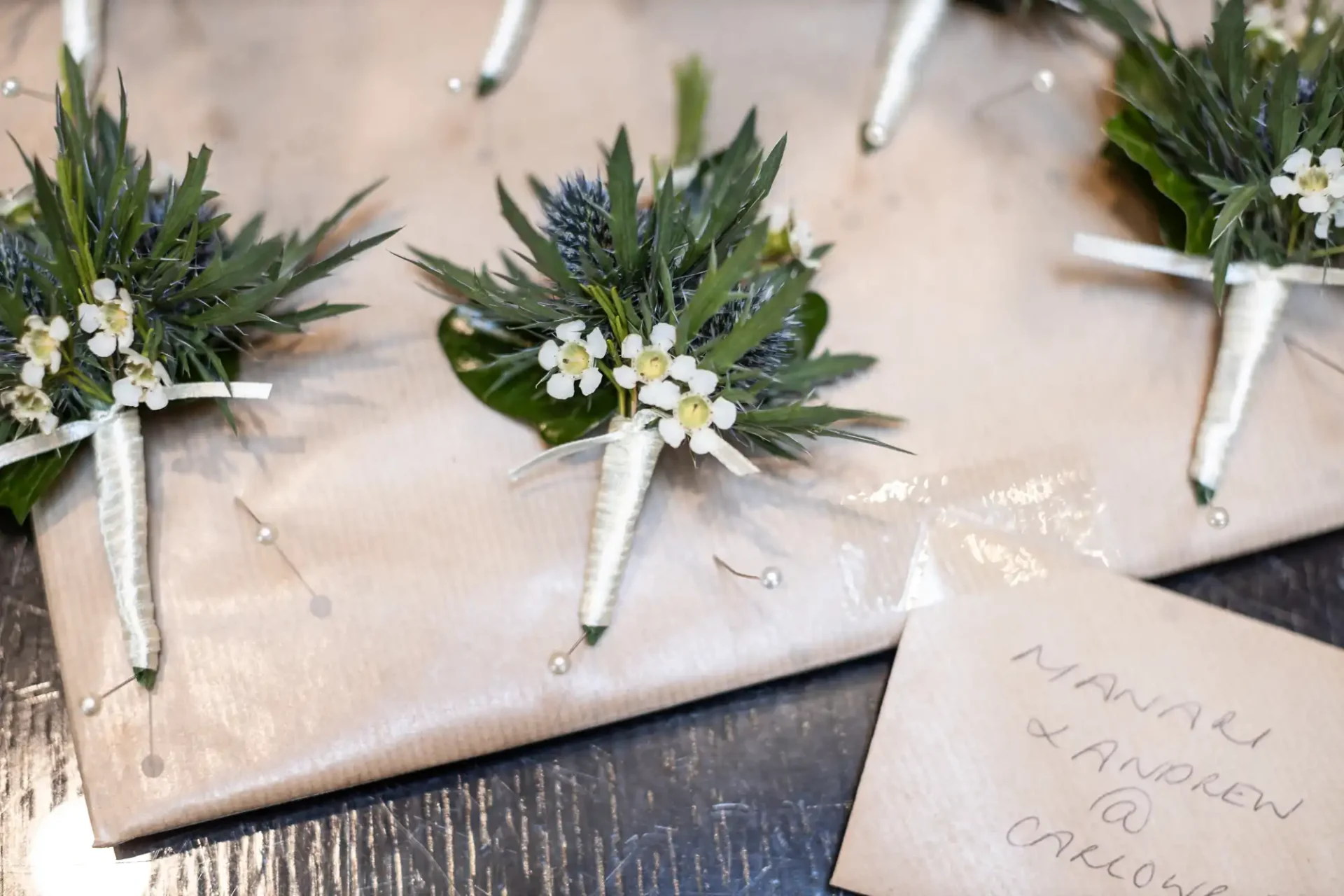 Boutonnieres with green leaves and white flowers are arranged on a wrapped surface. A card with handwritten text is partially visible.