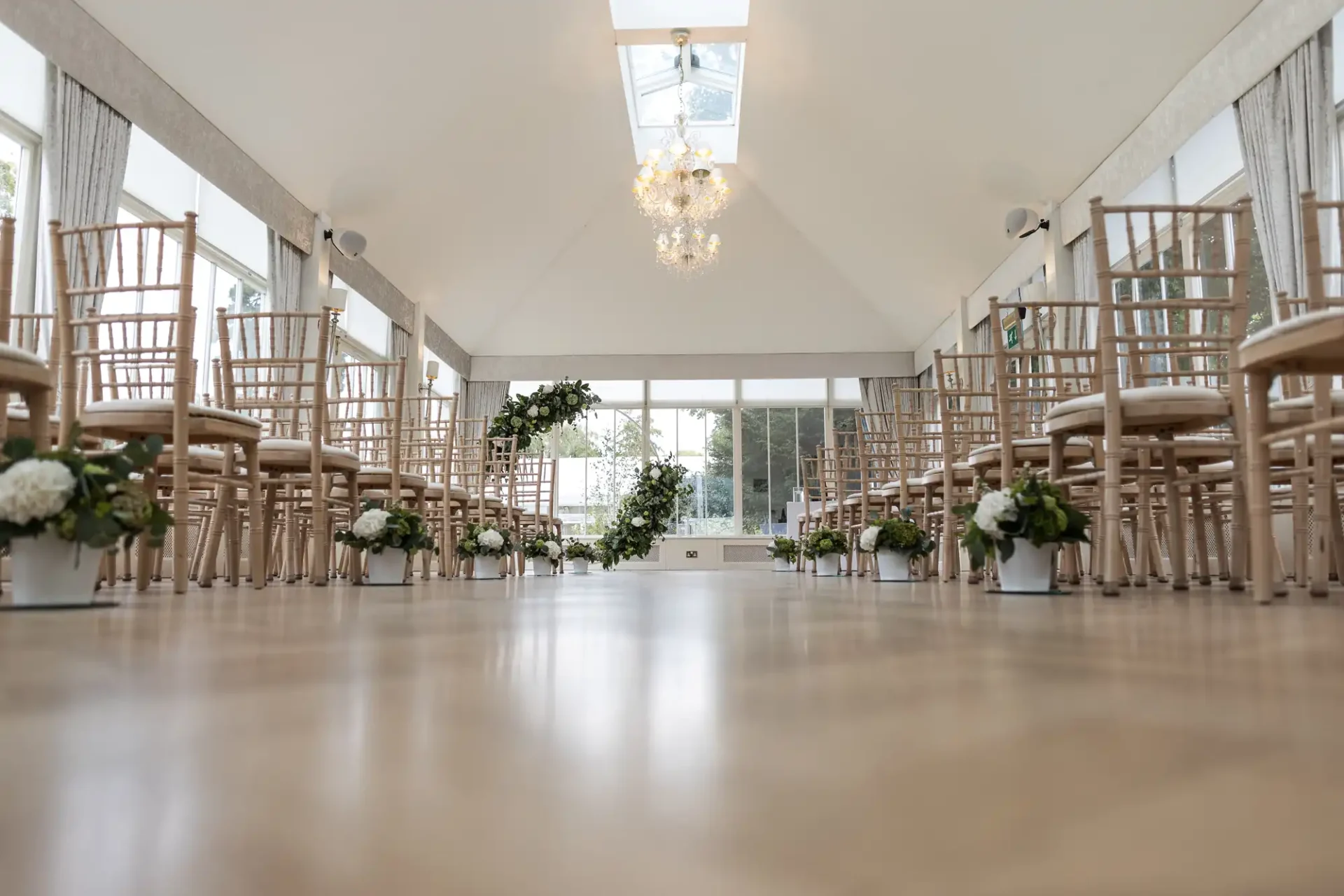 An empty light-filled wedding ceremony room with rows of wooden chairs facing a simple floral arch at the back.