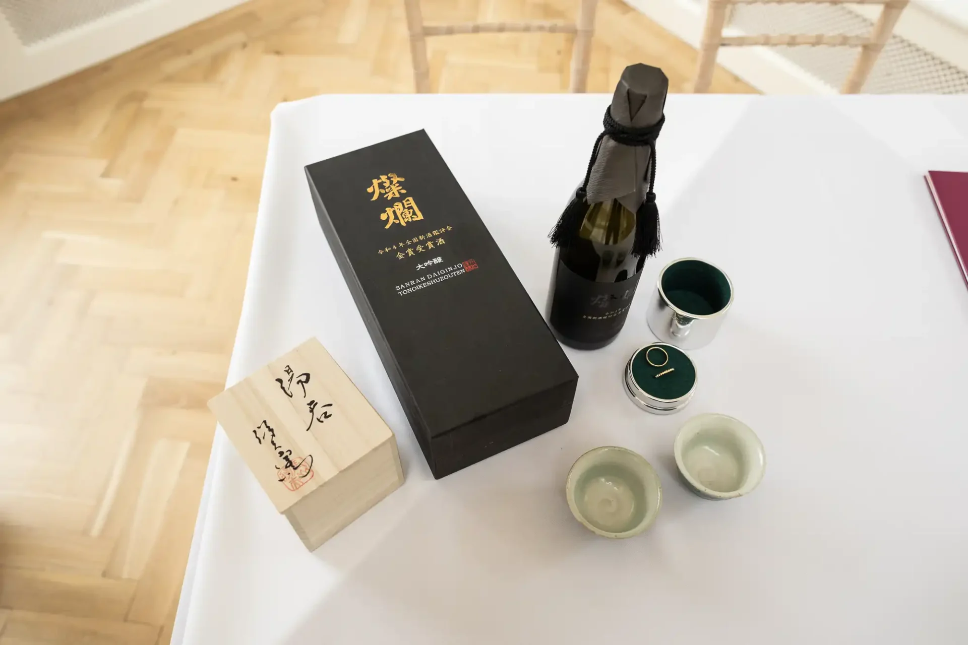 A boxed bottle with Japanese text, two ceramic cups, and a small wooden box with calligraphy sit on a white tablecloth.