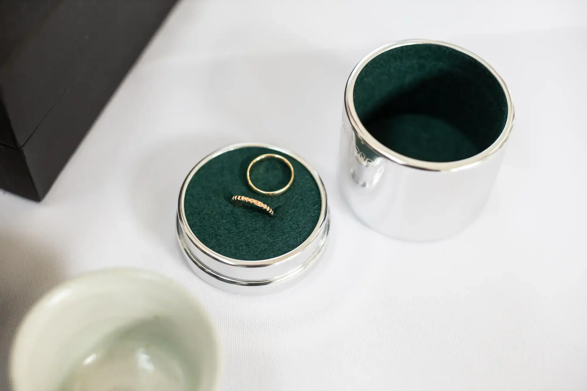 A silver jewelry container with a green interior holds a gold ring and a gold bracelet. The lid is placed nearby on a white surface.