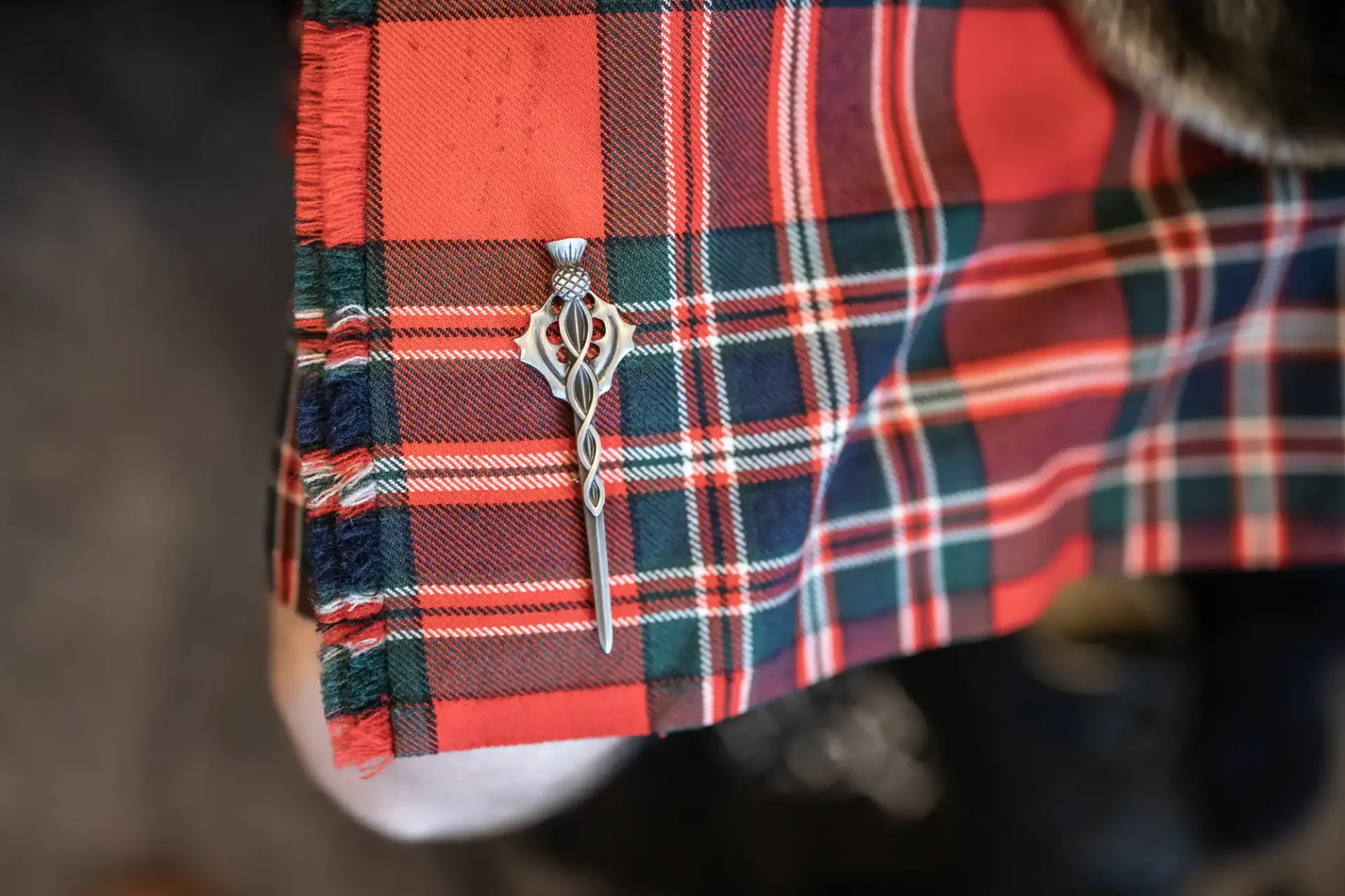 A silver kilt pin with a Celtic design is attached to a red, green, and blue tartan fabric.