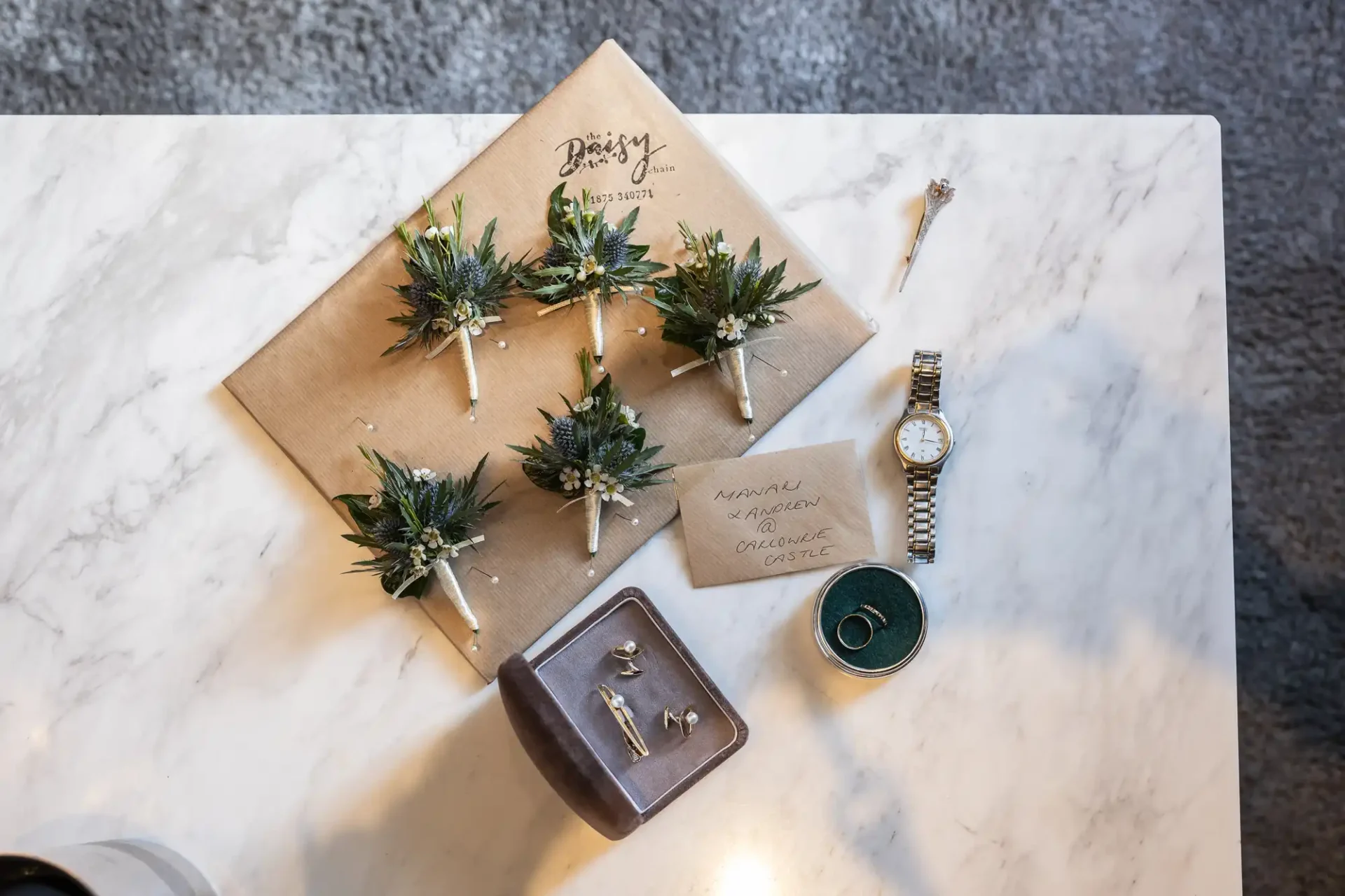 Boutonnieres on a marble table with a watch, wedding rings, note, and jewelry box.