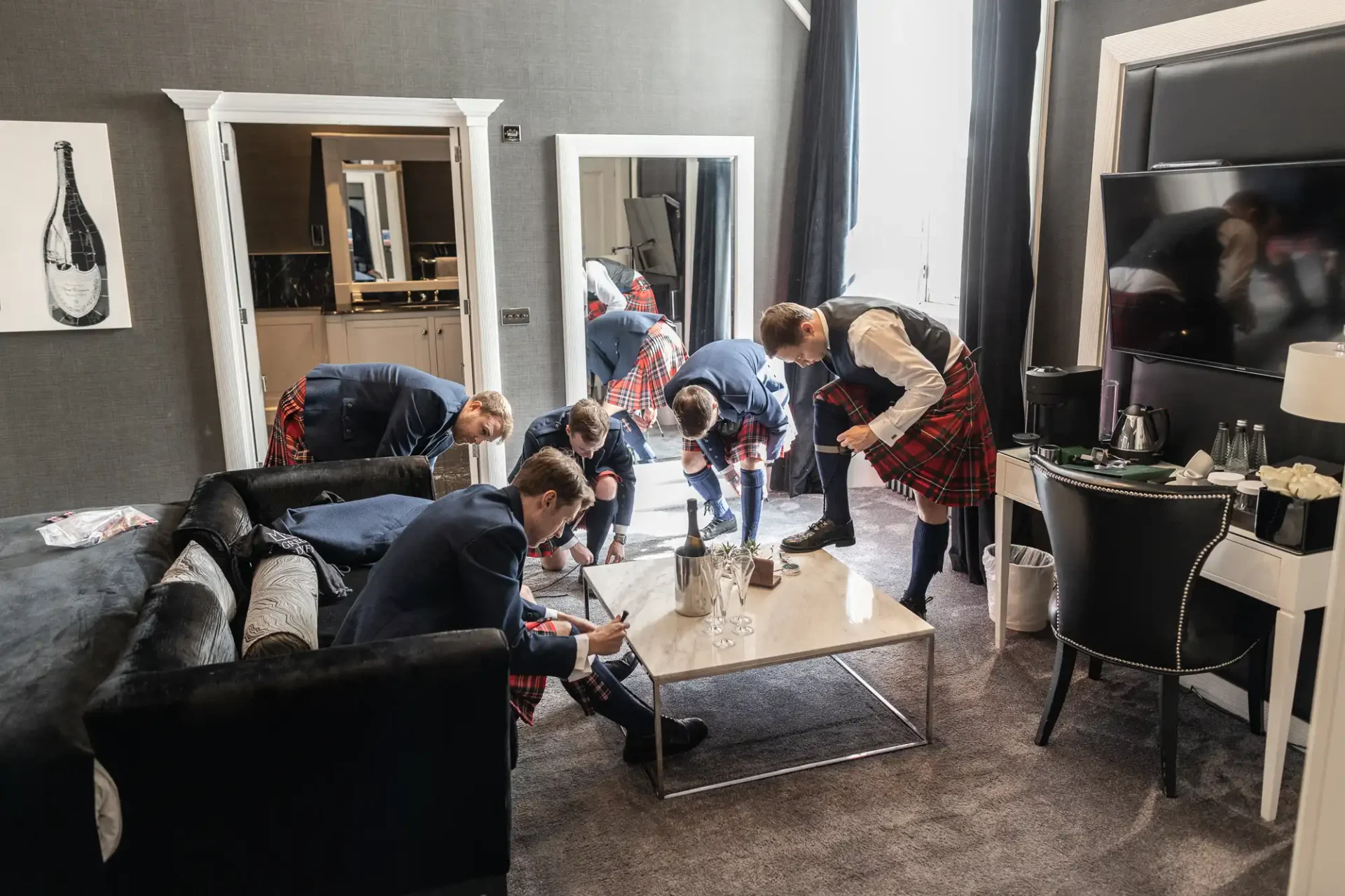 A group of men in kilts prepare for an event in a hotel room, focusing on tasks around a coffee table. The room is modern, with a sofa, TV, and wall art.