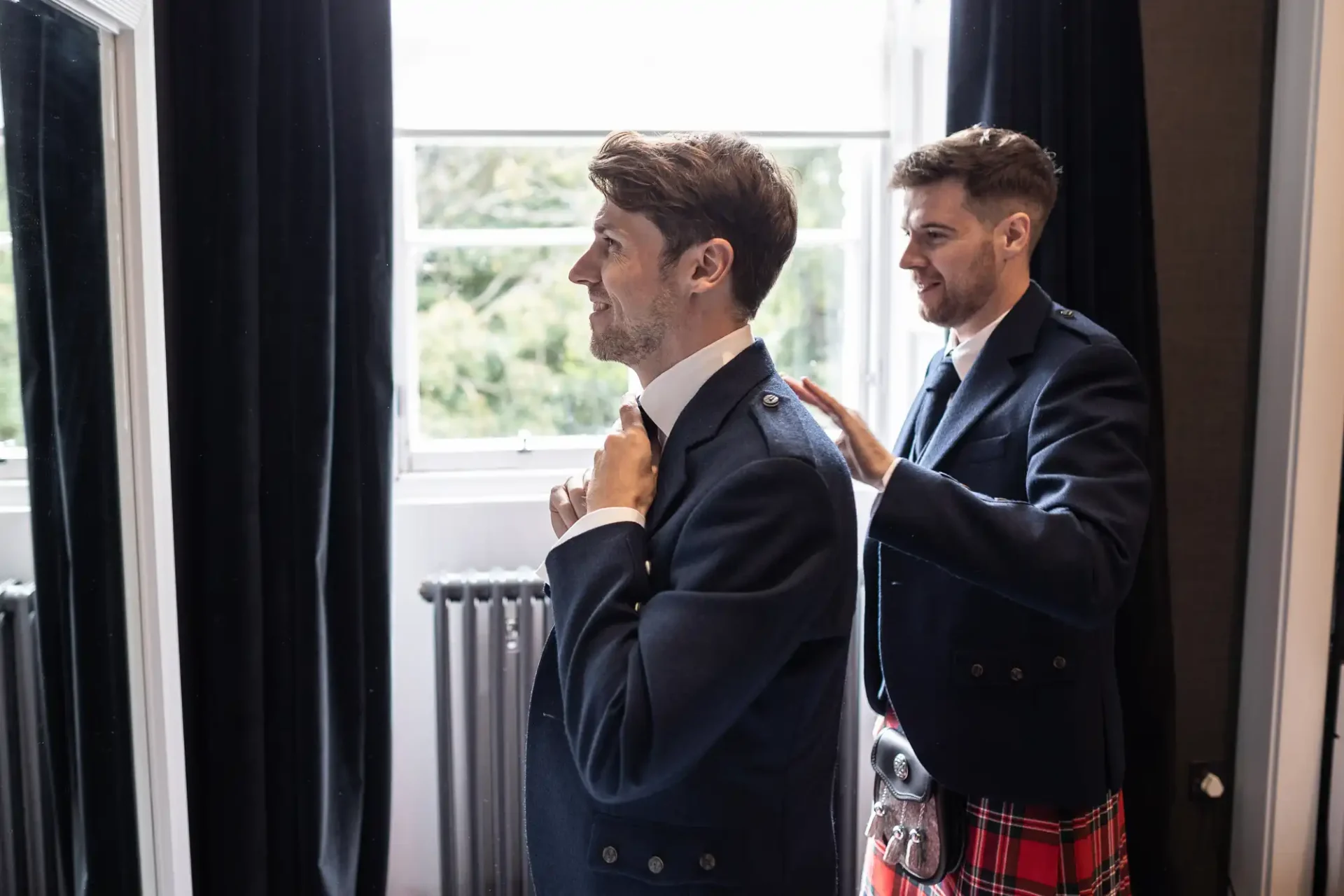 Two men in formal attire stand by a window. One man adjusts his collar while the other helps him with his jacket.