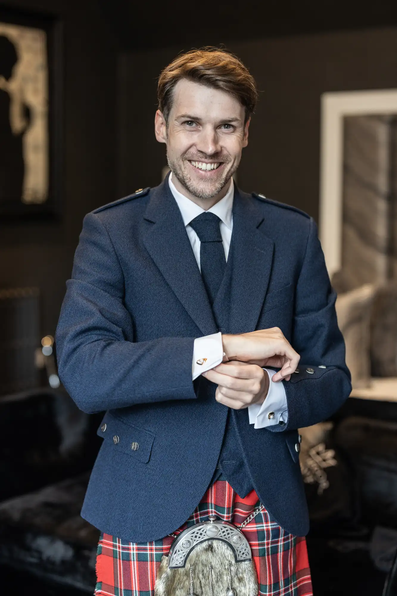 A man in a blue jacket and tie smiles while adjusting his cufflinks. He is wearing a red tartan kilt and standing in a room with dark walls and framed art.