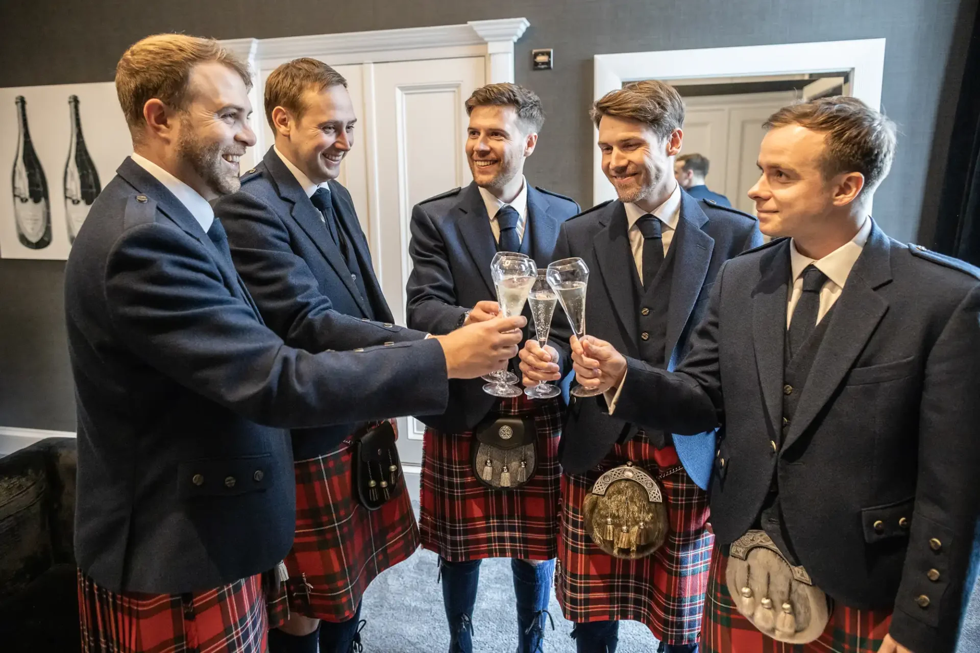 Five men wearing Scottish kilts and jackets raise glasses in a toast, standing in a room with framed artwork on the wall.