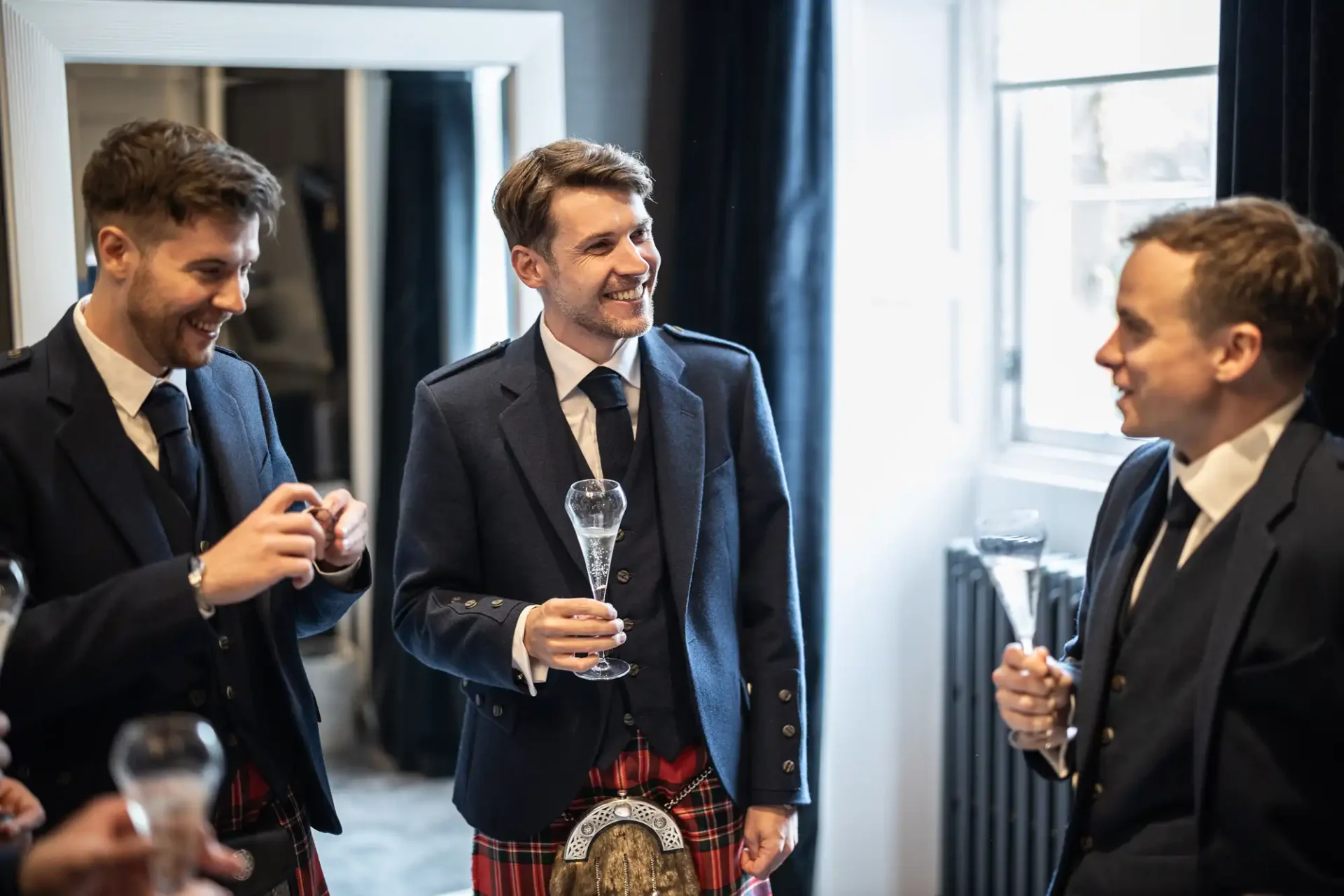 Three men in suits with tartan kilts stand indoors holding glasses. They are smiling and engaged in conversation.