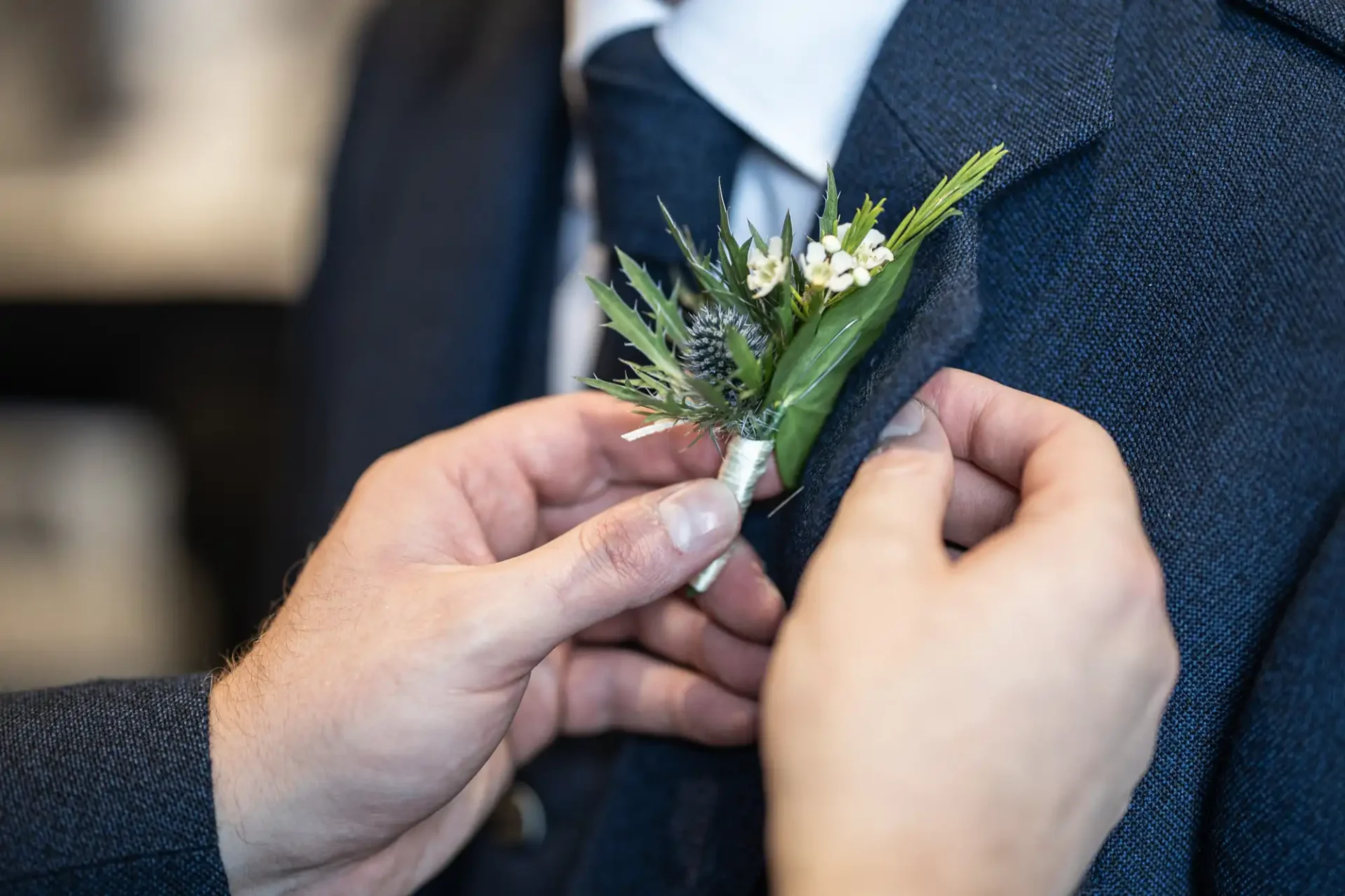 Person attaching a boutonniere with greenery and small white flowers to a man's dark suit jacket.