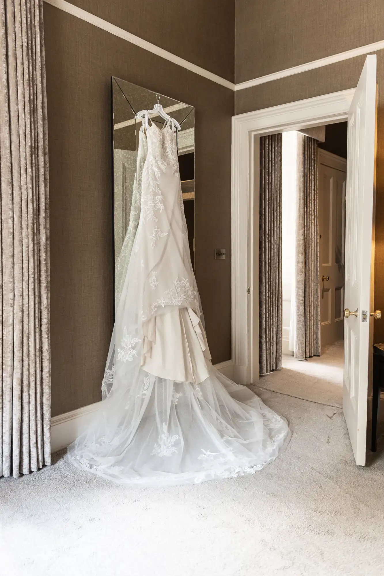 A white lace wedding dress hangs on a mirror against a beige wall in a carpeted room with an open door.