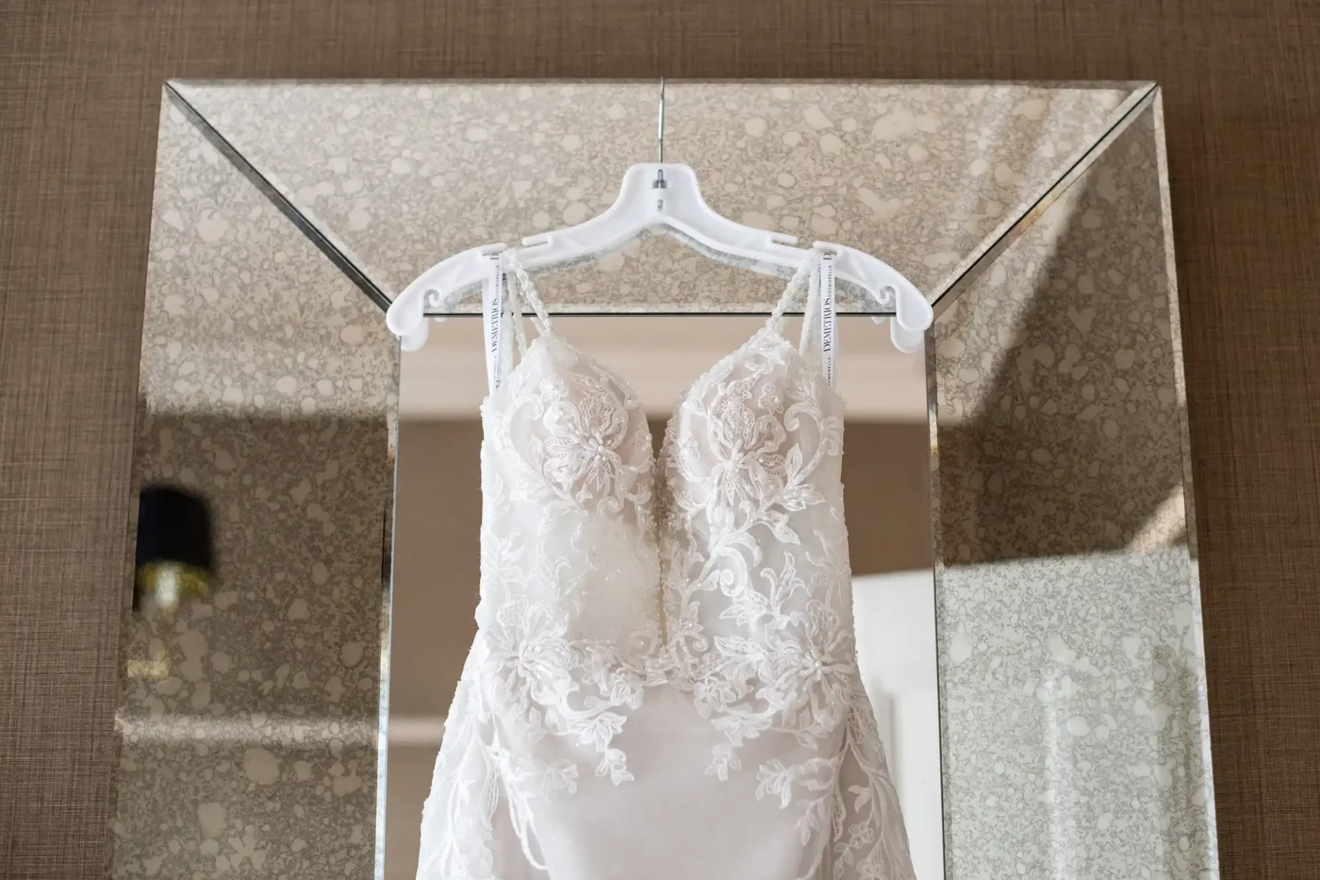 A white lace wedding dress hangs on a hanger in front of a large rectangular mirror.