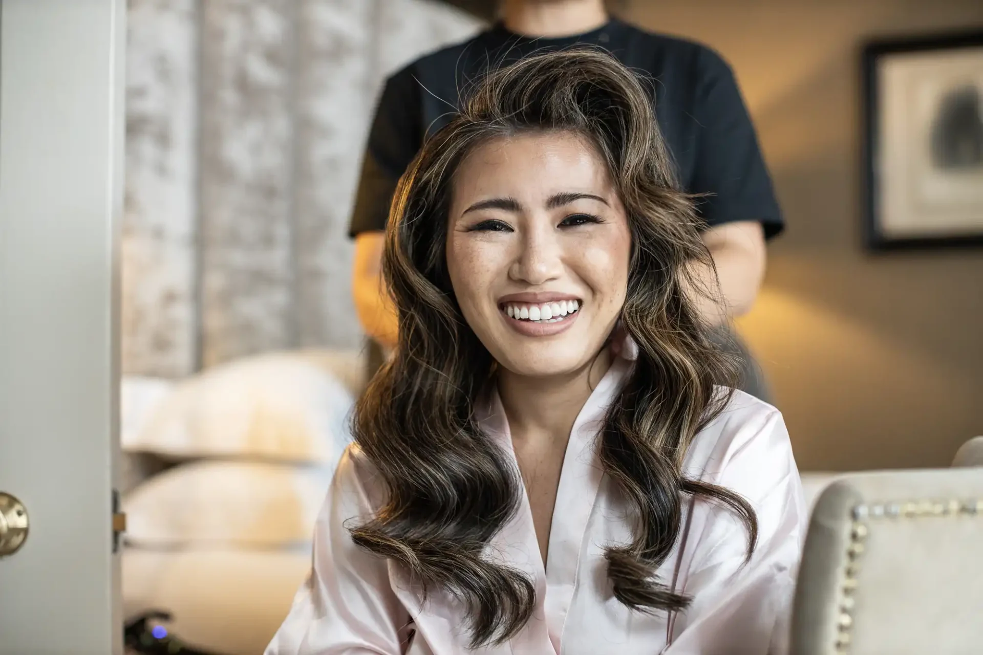 Smiling person with long wavy hair wearing a light-colored robe, sitting indoors.