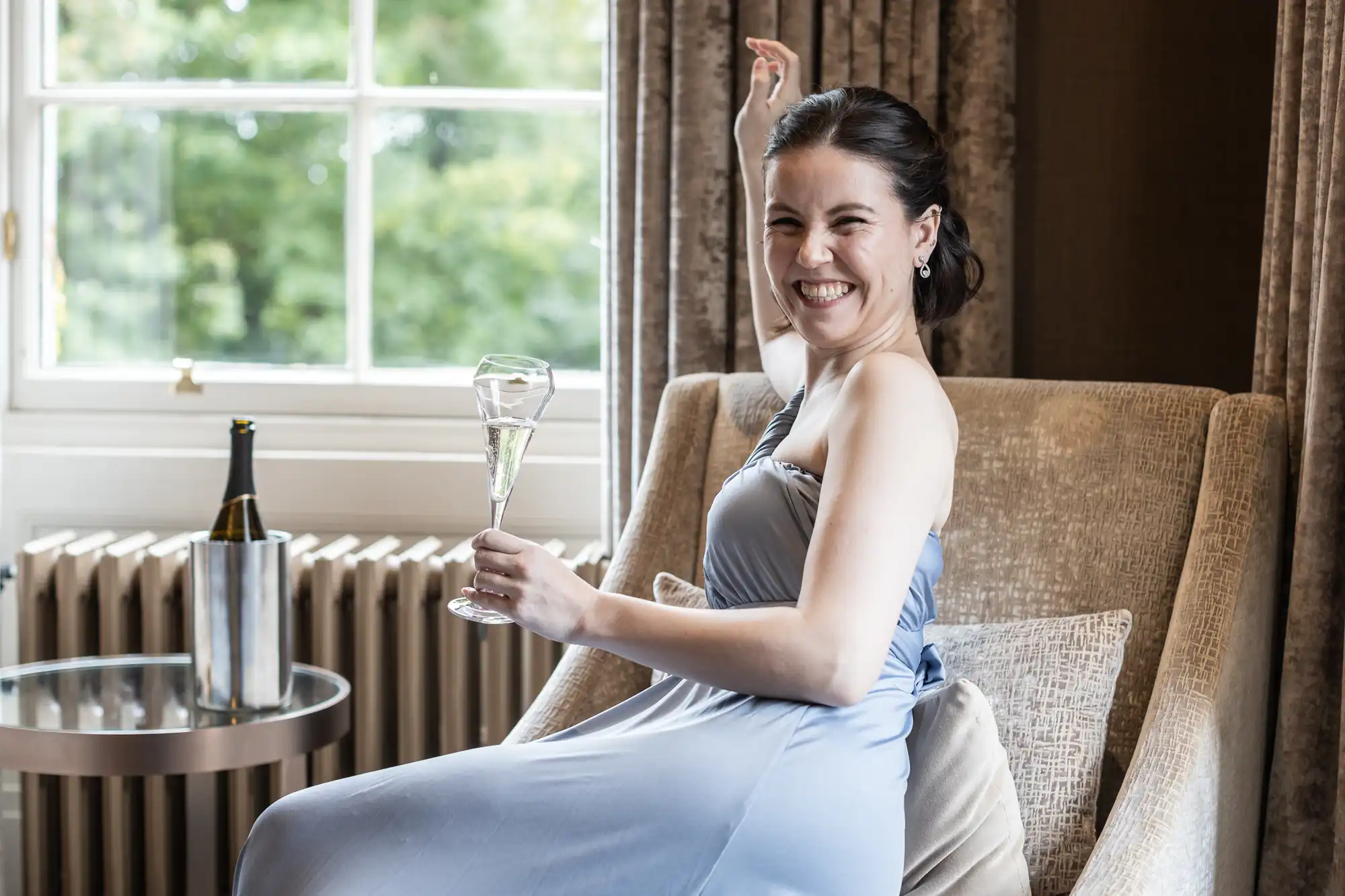 A woman in a gray dress smiles while holding a champagne glass, sitting on a chair near a window with a champagne bottle on a side table.