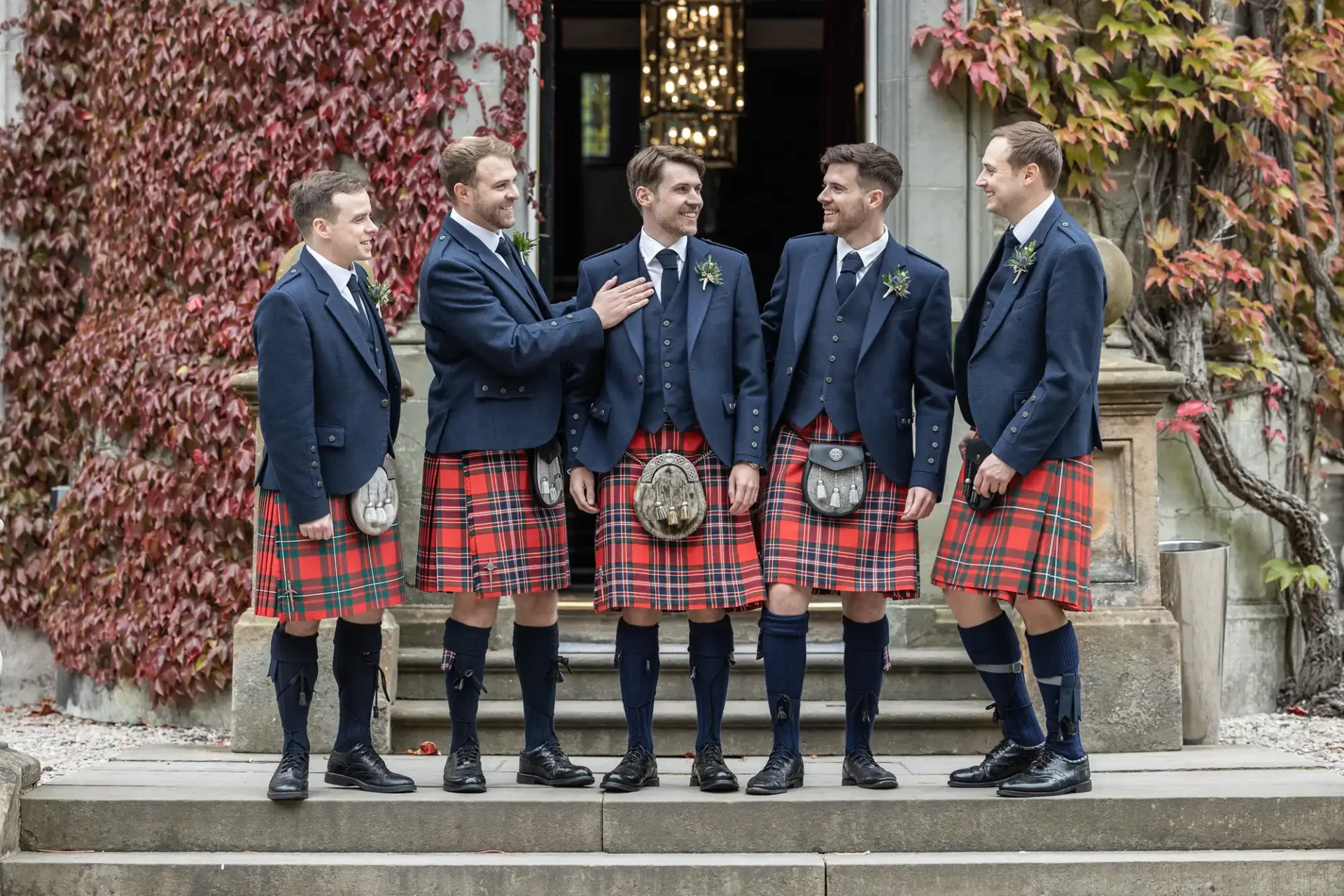 Five men in matching blue jackets and red tartan kilts stand together on stone steps, surrounded by ivy-covered walls.