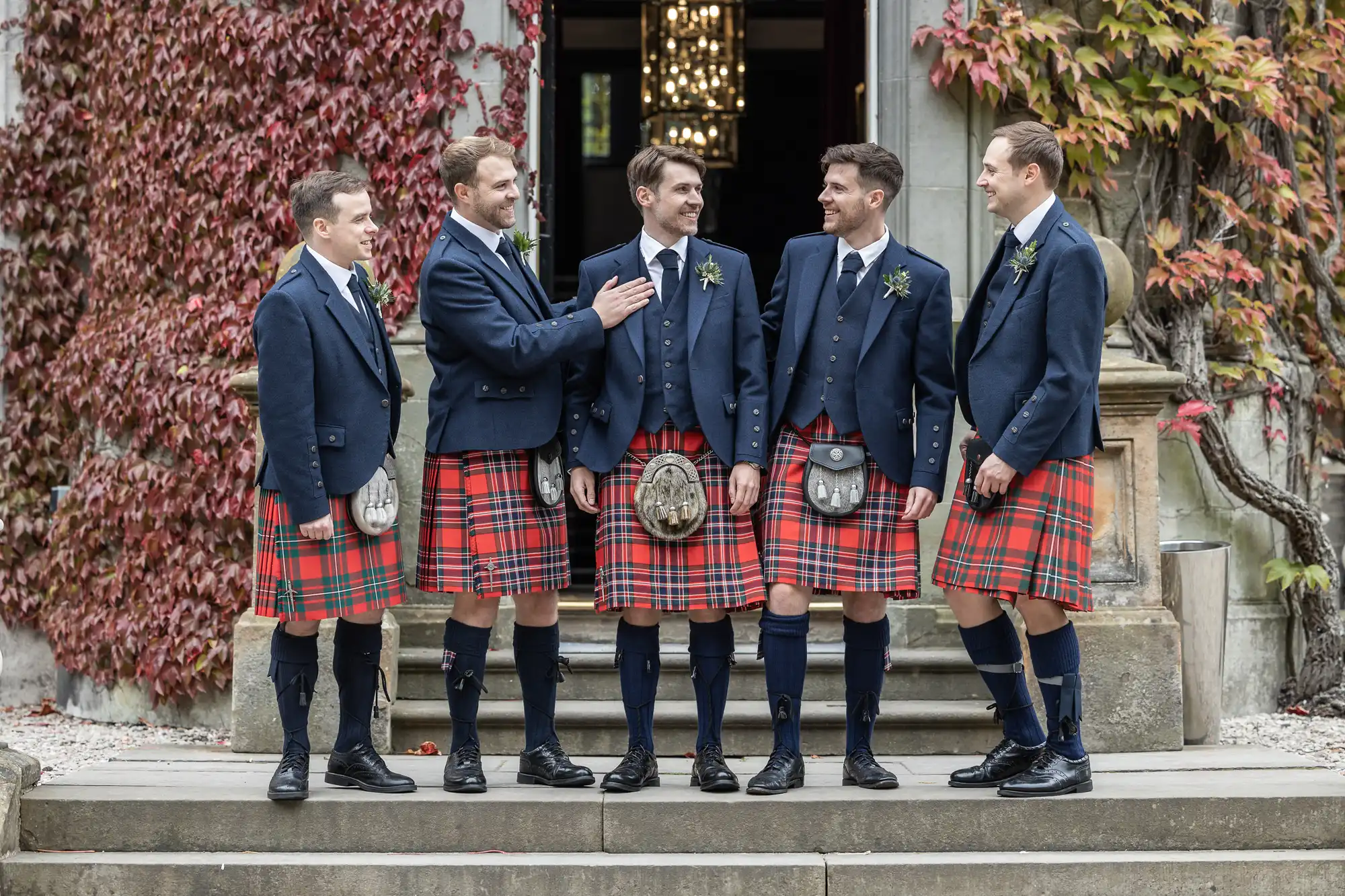 Five men in matching blue jackets and red tartan kilts stand together on stone steps, surrounded by ivy-covered walls.