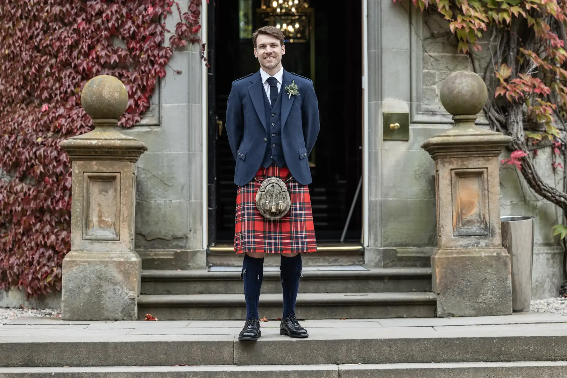 Man in a blue jacket and red tartan kilt stands on stone steps in front of an ivy-covered entrance.