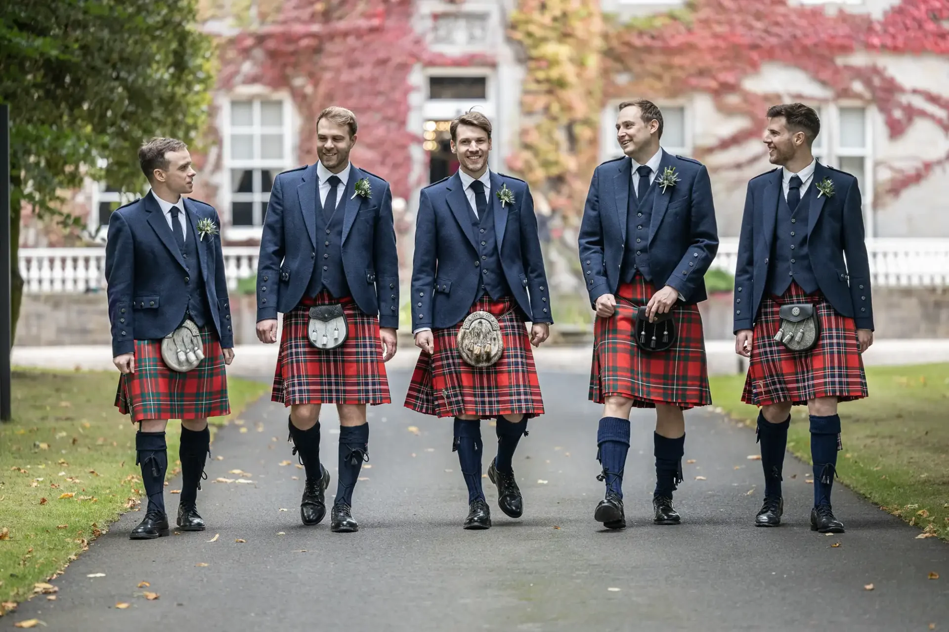 Five men wearing blue jackets and red tartan kilts walk on a path, smiling and talking.