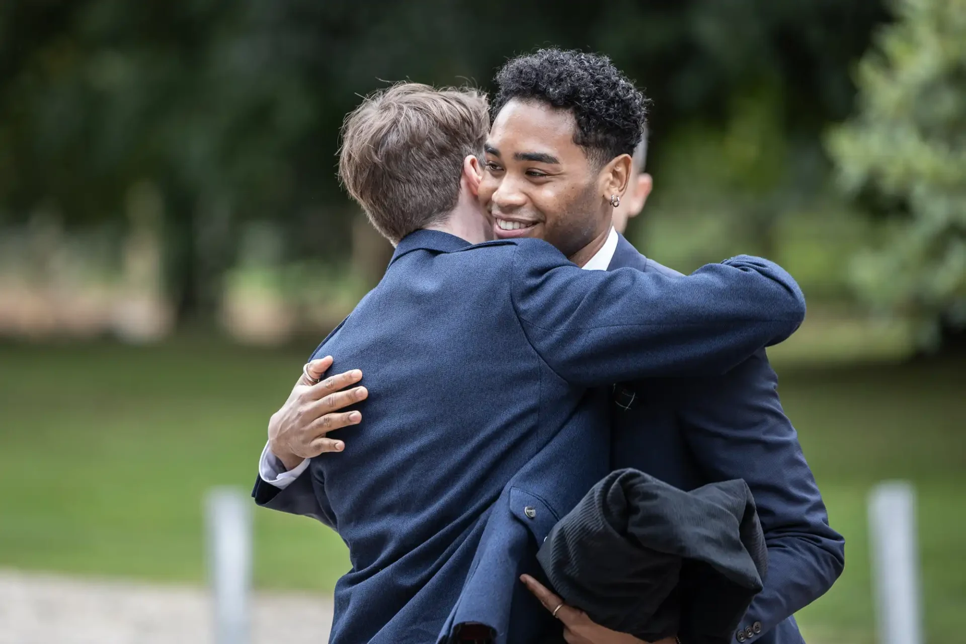 Two men in suits hugging warmly outdoors with greenery in the background.