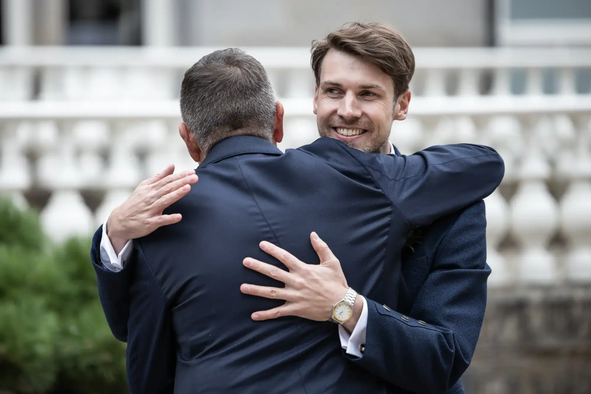 Two men in suits hugging outdoors, with a blurred background of a white balustrade. The man facing the camera is smiling.