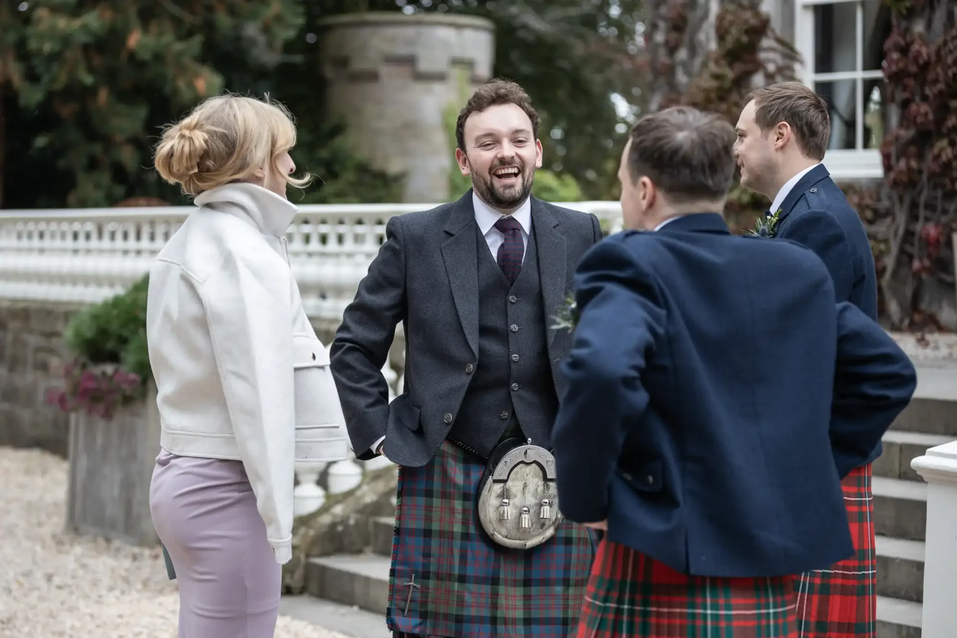 A group of people in formal attire, including two men wearing kilts, stand outside a building. One man is smiling and talking to a woman with blonde hair.