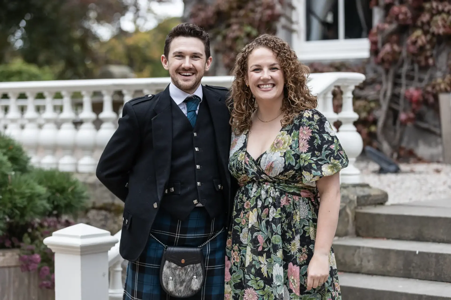 A man in a traditional Scottish outfit stands next to a woman in a floral dress, both smiling, in front of a white railing and stone steps.