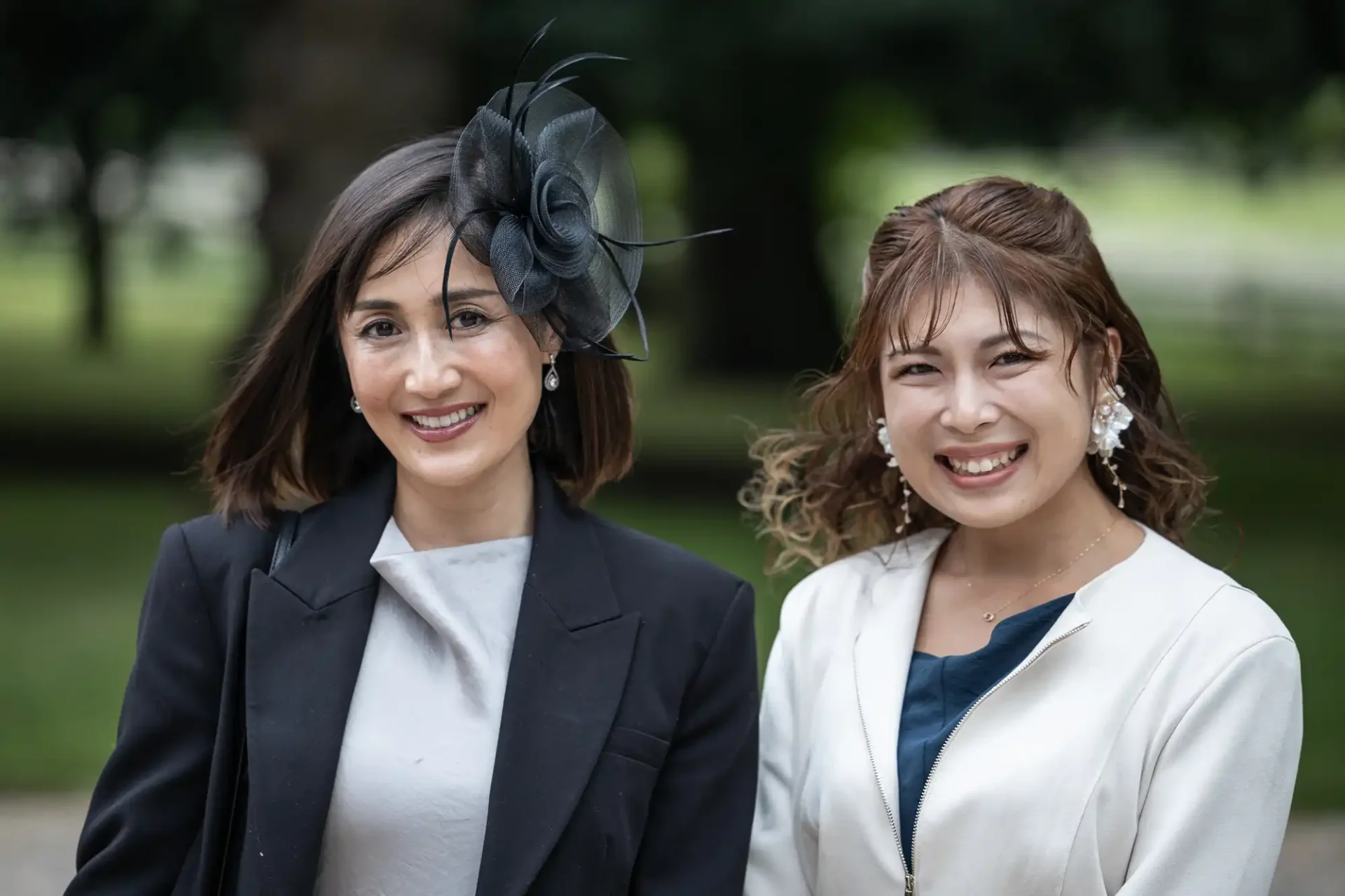 Two women smiling, one wearing a black fascinator and black blazer, the other in a light-colored blazer. Greenery in the background.