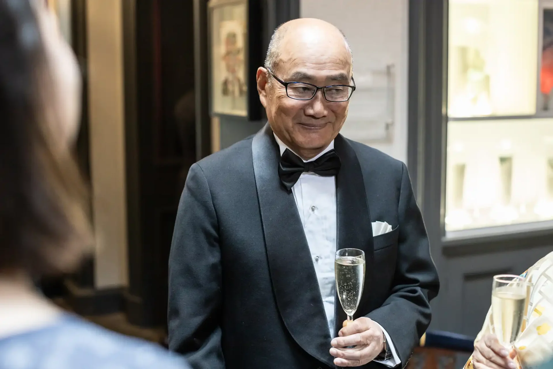 An older man in a tuxedo holds a glass of champagne, engaging with others at a formal event.