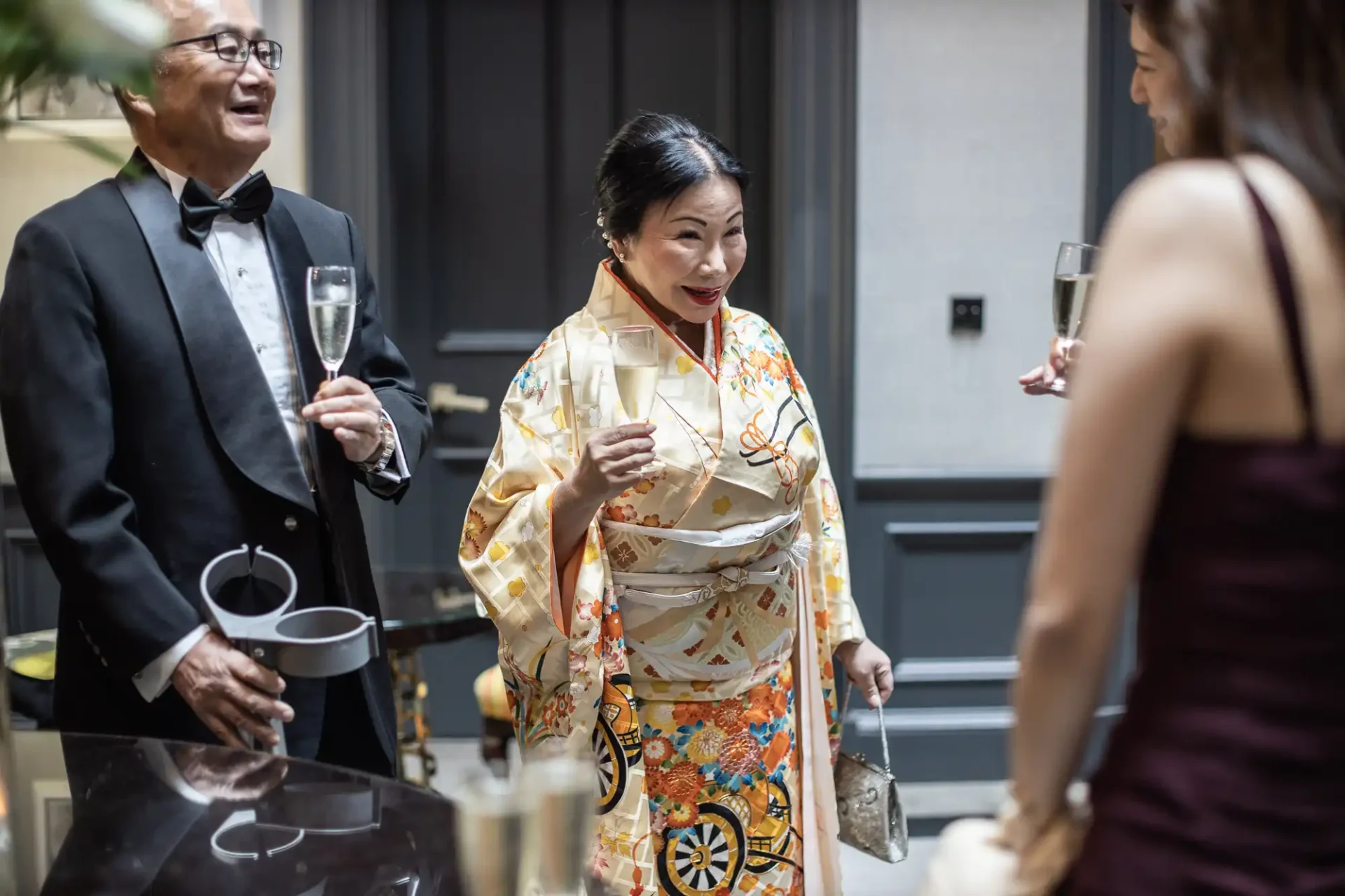 A woman in a colorful kimono holds a champagne glass and smiles in conversation with a man in a tuxedo and a woman in a dark dress at an indoor gathering.