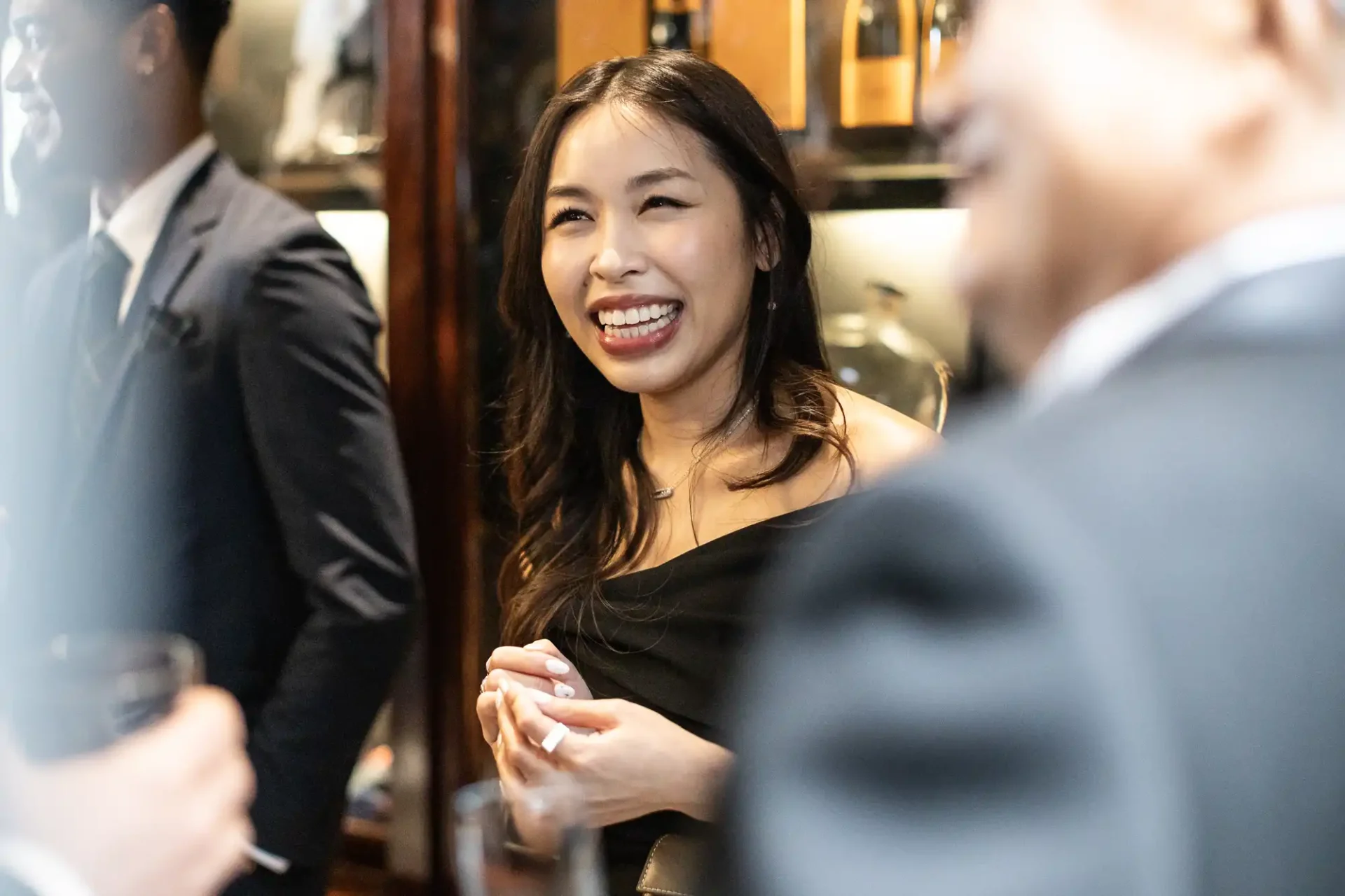 A woman in a black dress is smiling and holding a glass in a social gathering with three other people in suits.