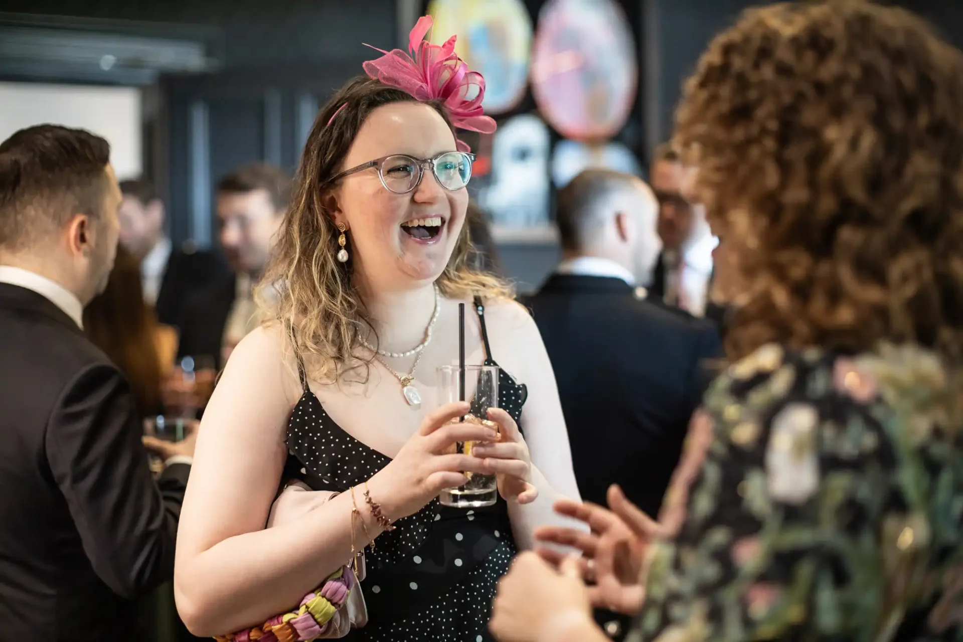 A woman in a polka dot dress, wearing glasses and a pink fascinator, holding a drink and smiling while talking to another person at a social event.