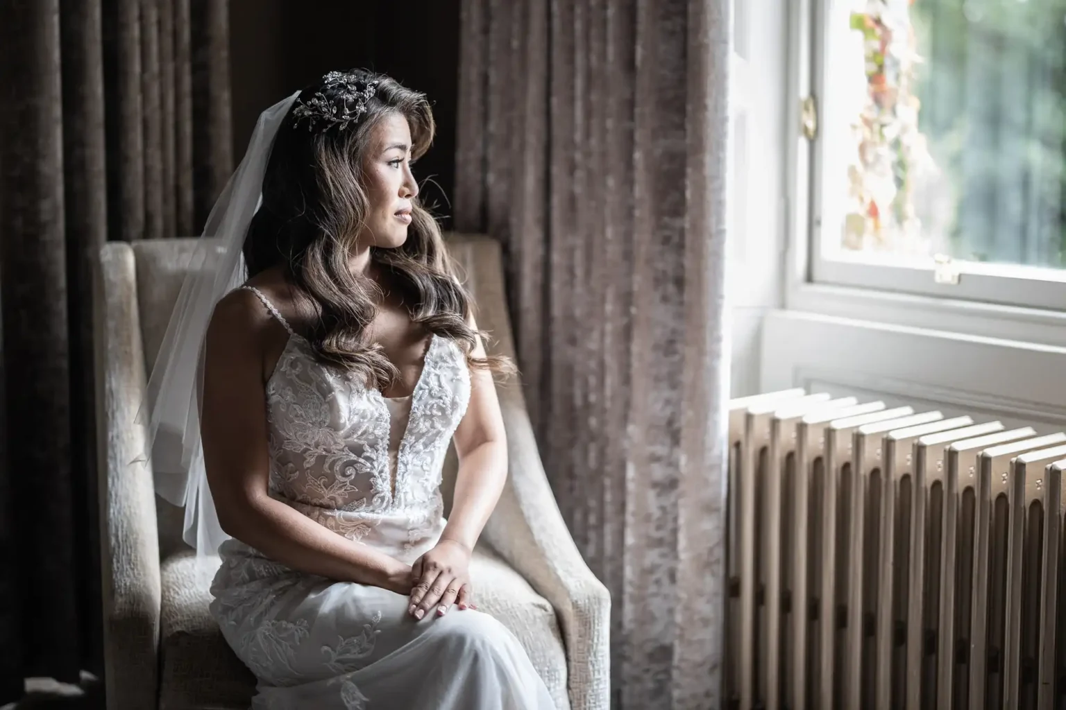 A bride in a white lace dress sits in a chair by a window, looking outside. She wears a floral headpiece with a veil.