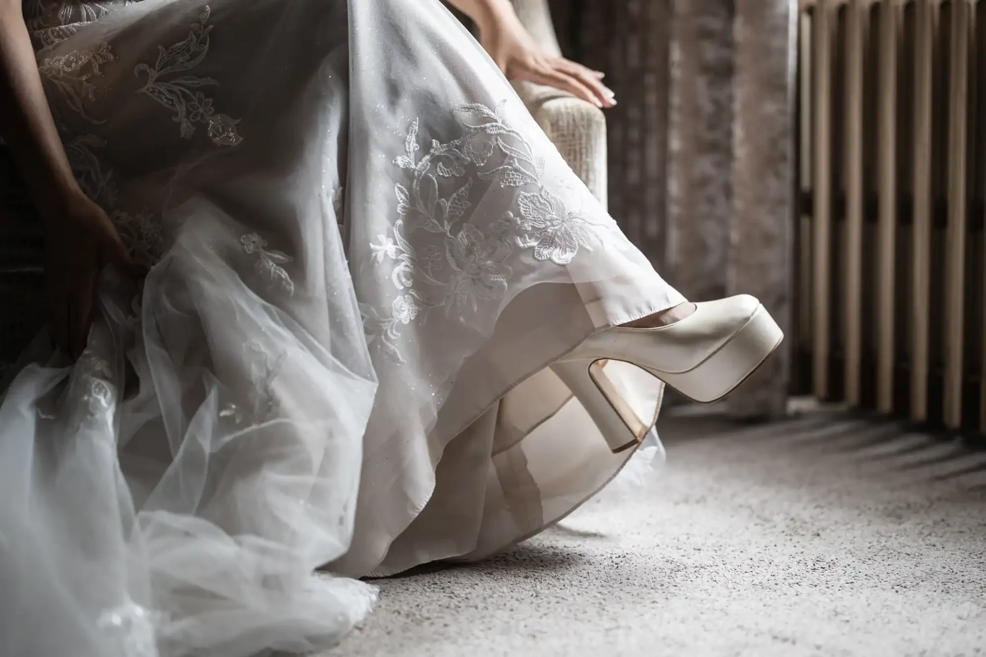 A person in a white lace wedding dress and high heels sits on a chair, with sunlight streaming through a window.