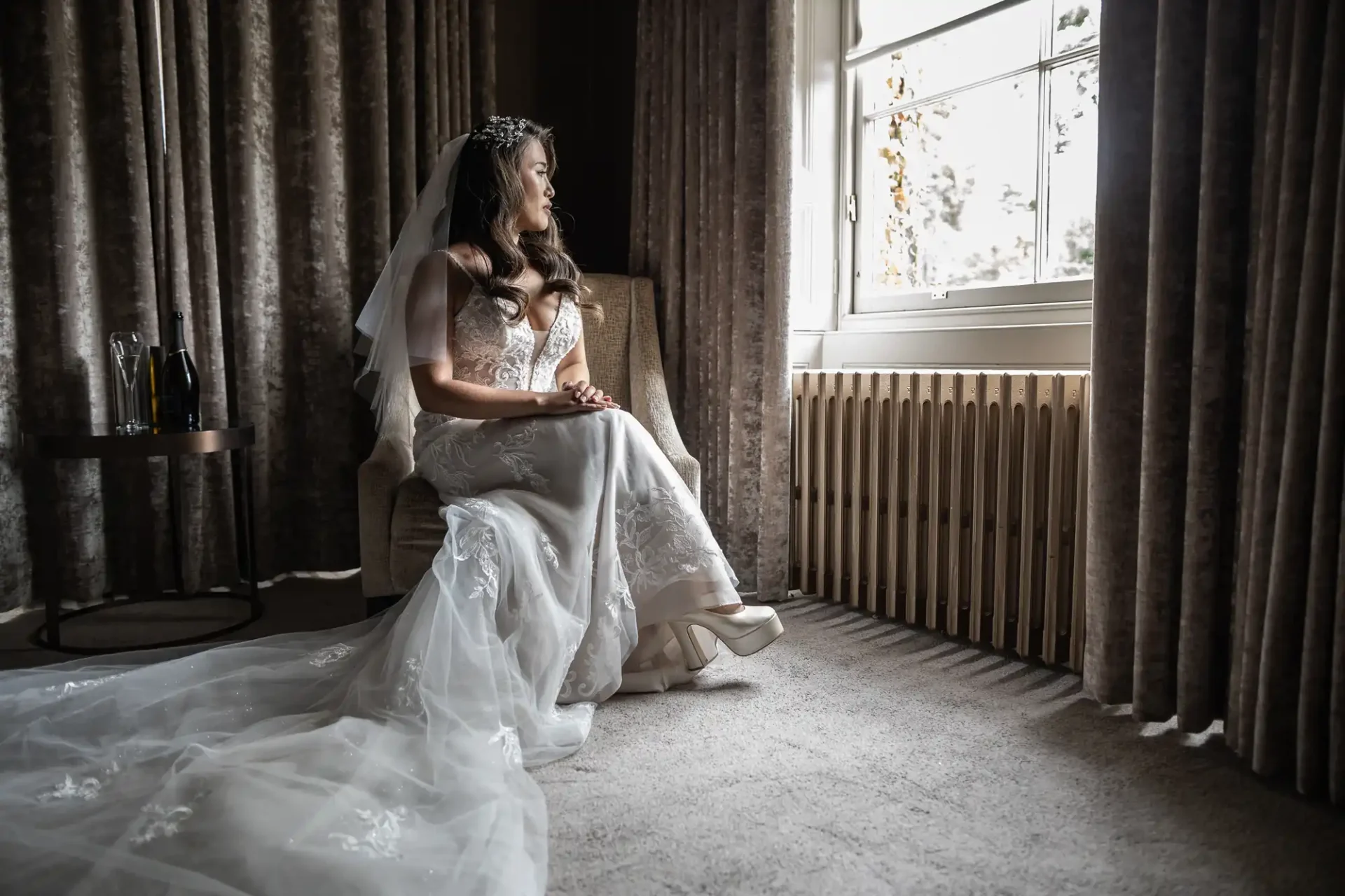 Bride in a white gown sits on a chair, looking out a window. She has a veil and tiara, and her hands are on her lap. A small table with a bottle is beside her.