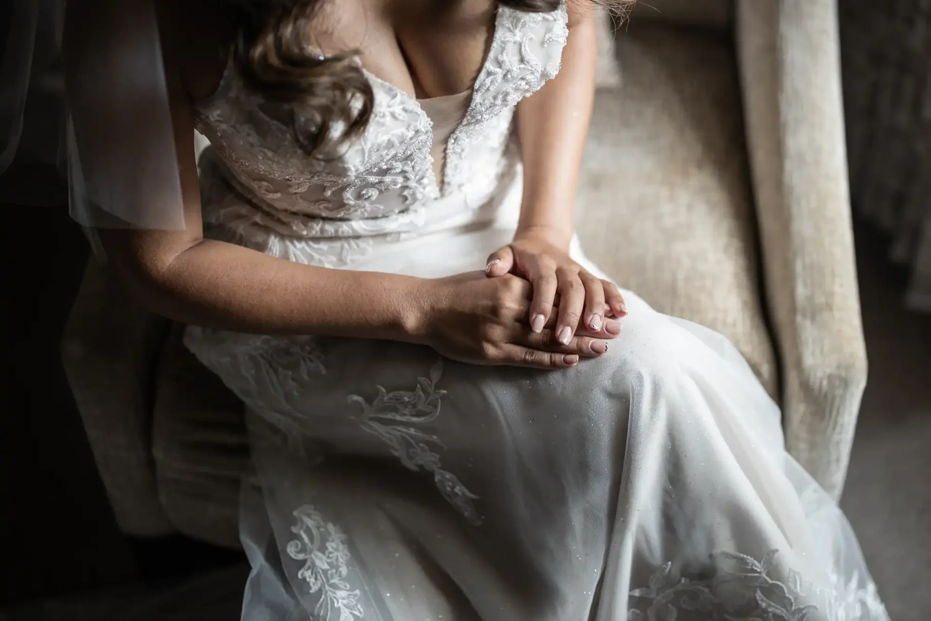 A bride in a white lace wedding dress sits on a chair, hands clasped on her lap, with her veil partially visible.