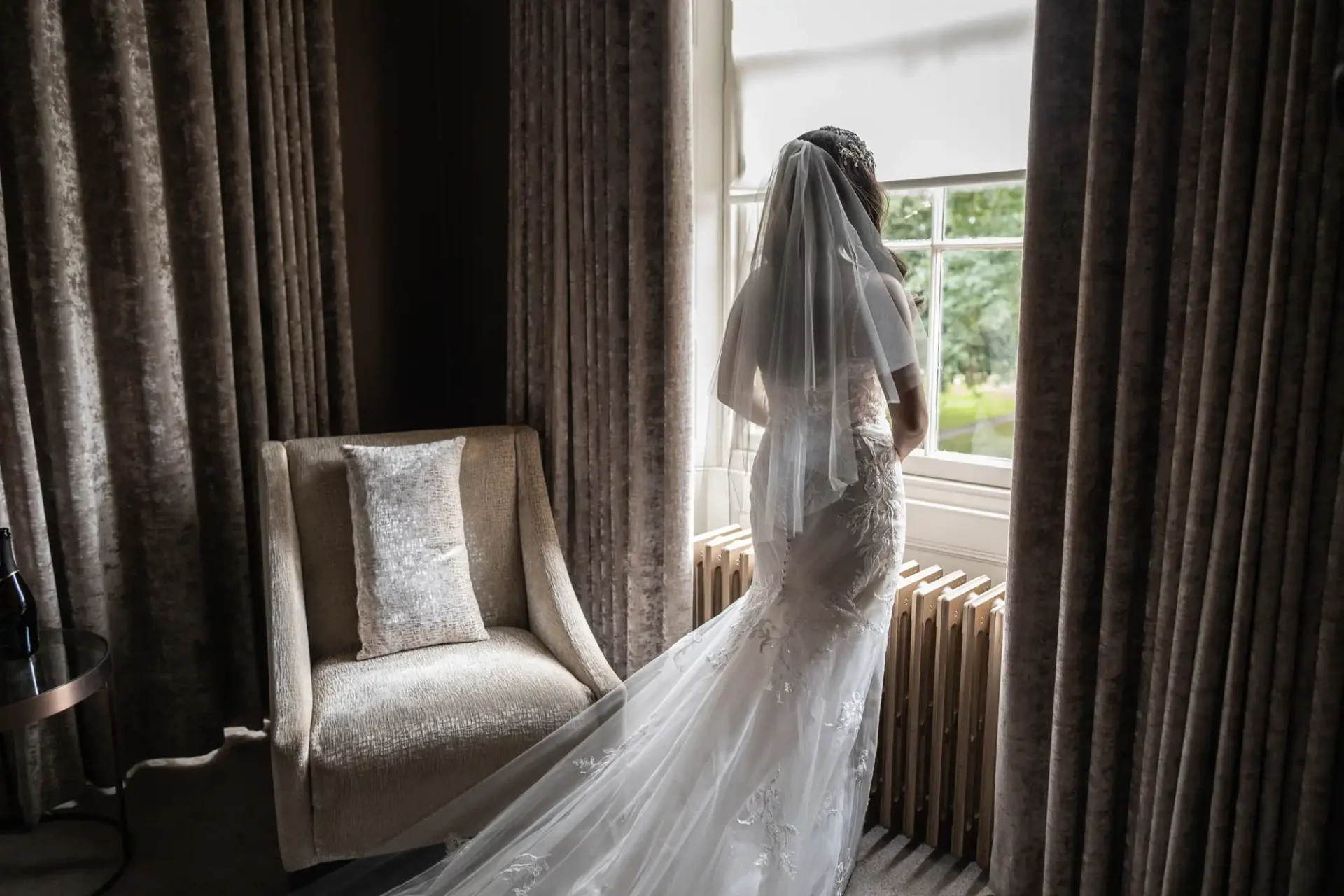 Bride in a white gown and veil stands by a window, looking outside. A cushioned chair and table with a bottle are by her side, with curtain-draped walls around.