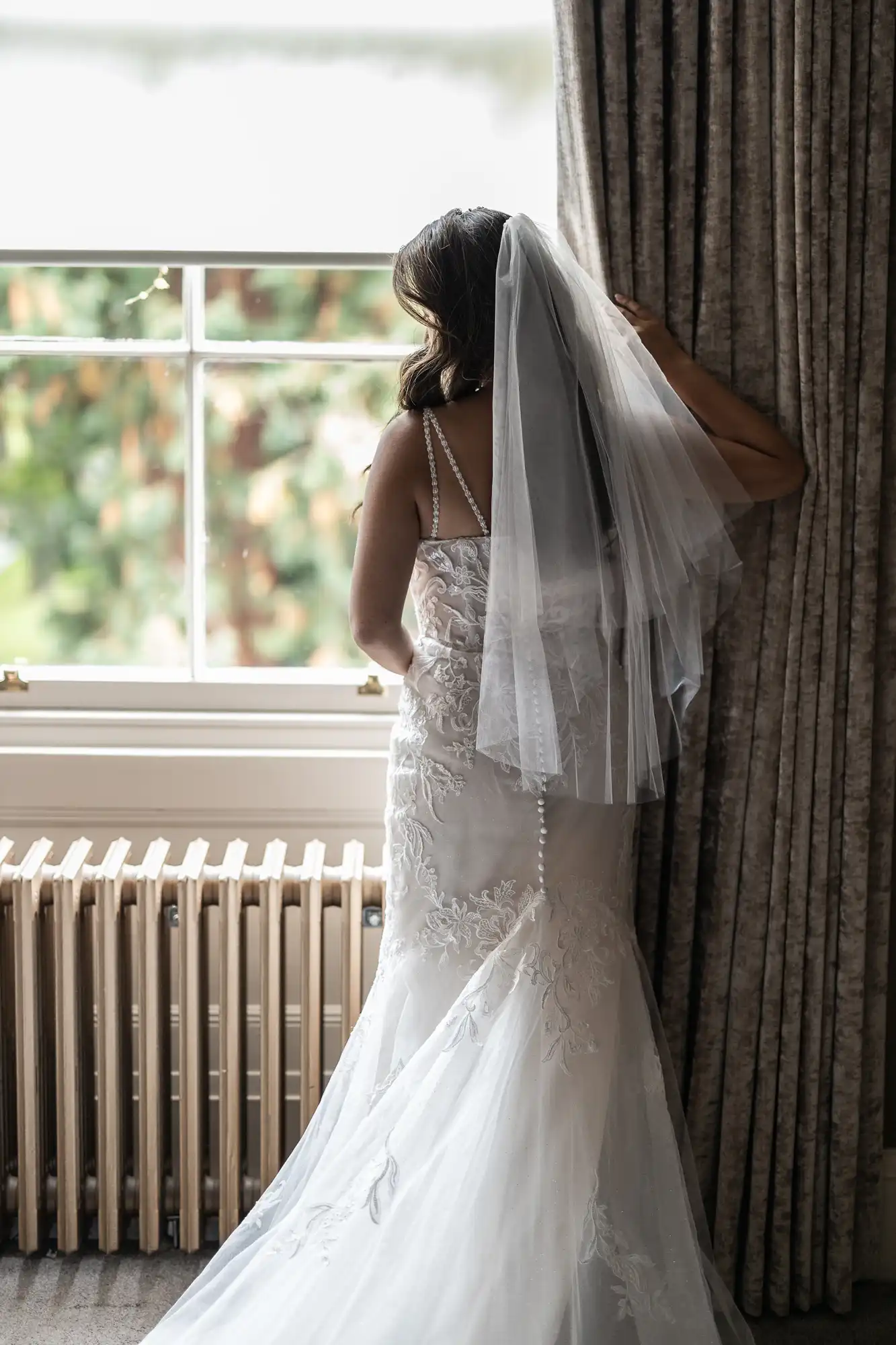 Bride in a white dress with veil, facing a window with greenery view, holding curtain.